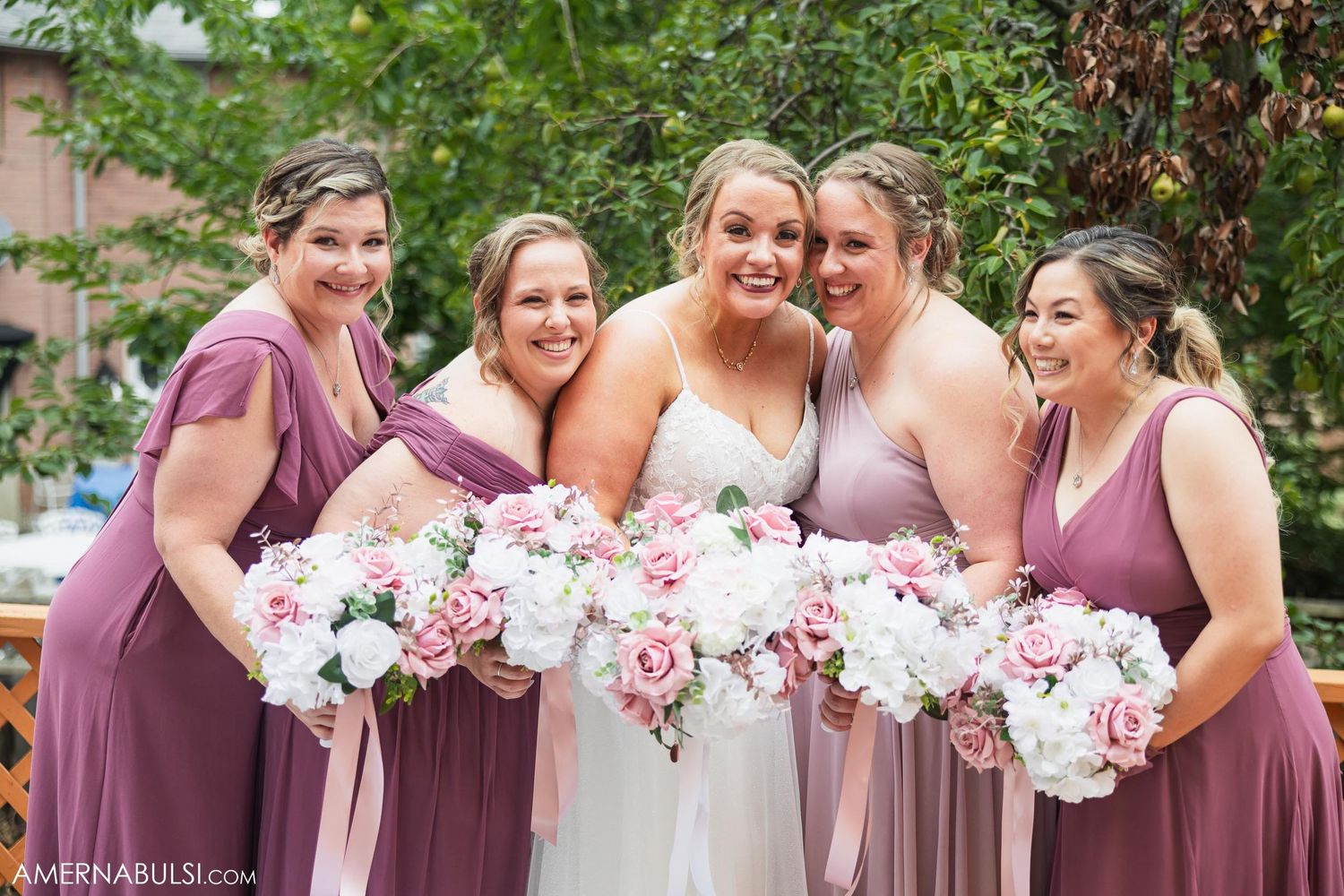 Bridesmaids in mauve dresses share joyful moment with white and pink bouquets.