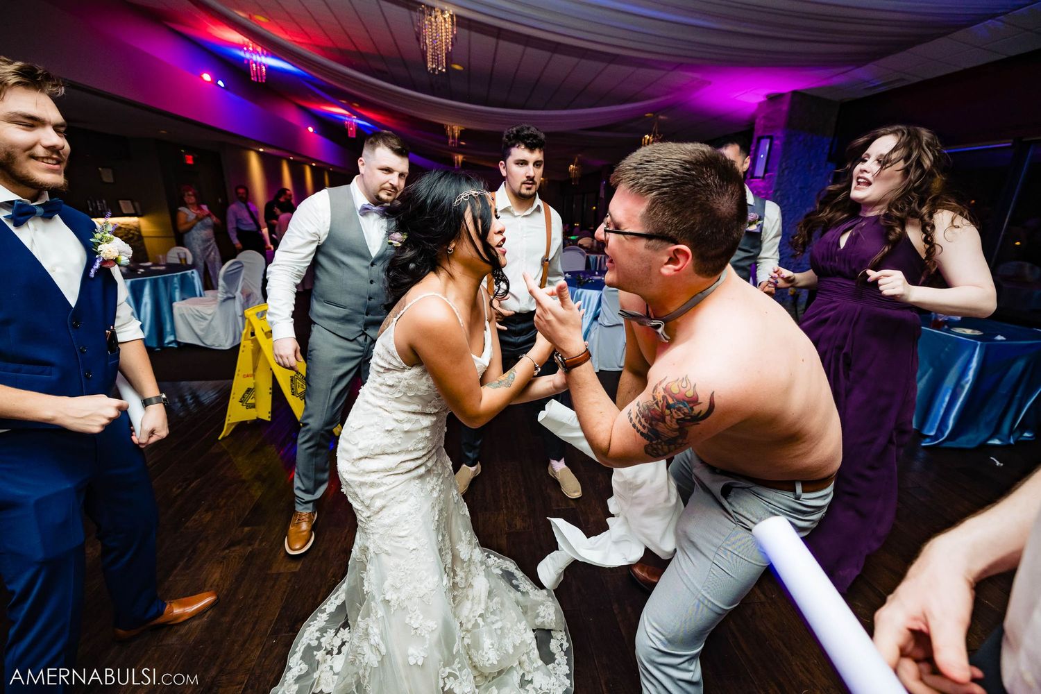 Wedding party dancing enthusiastically on wooden dance floor with purple lighting.