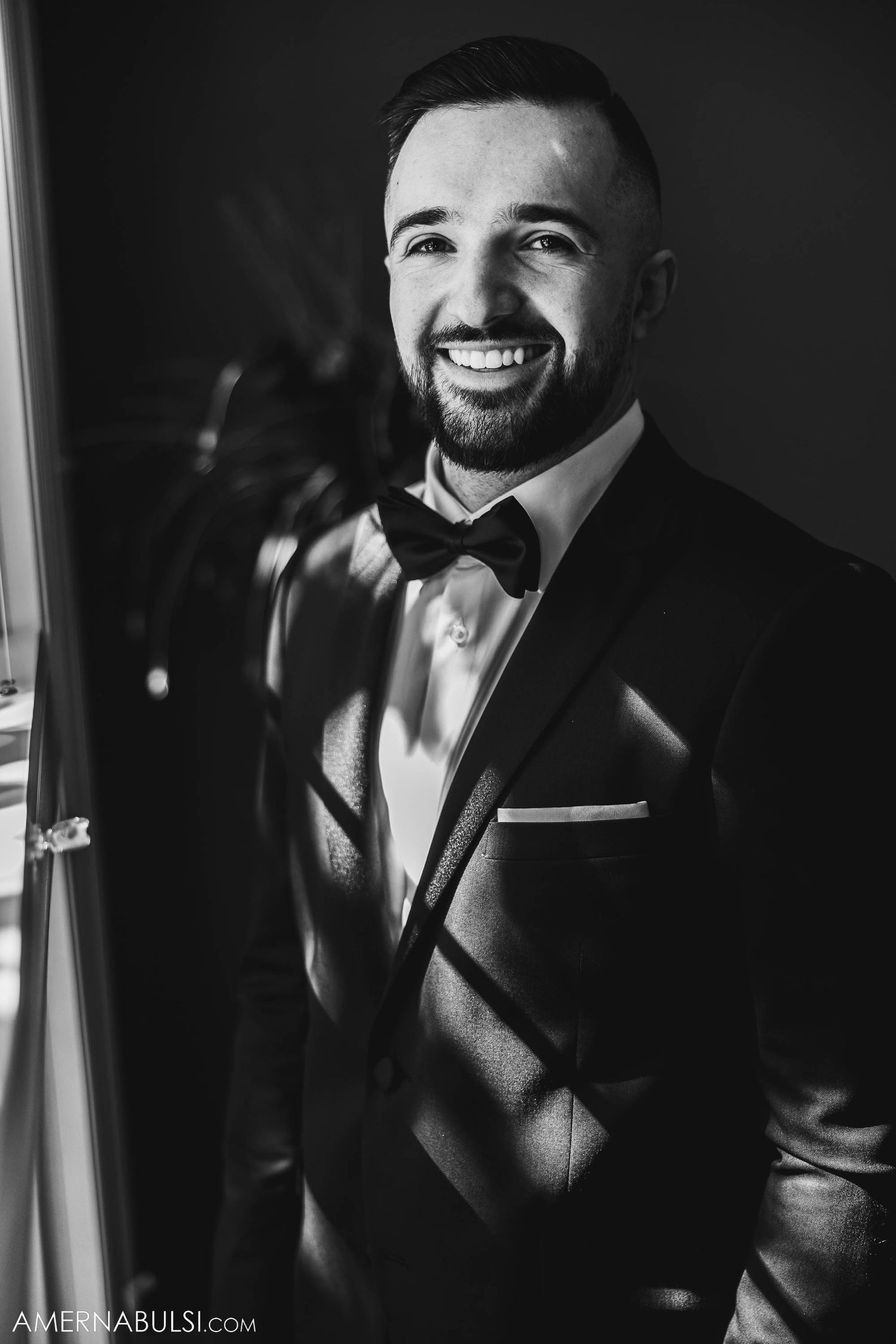 A black and white portrait of a groom in a tuxedo standing by a window with dramatic lighting.
