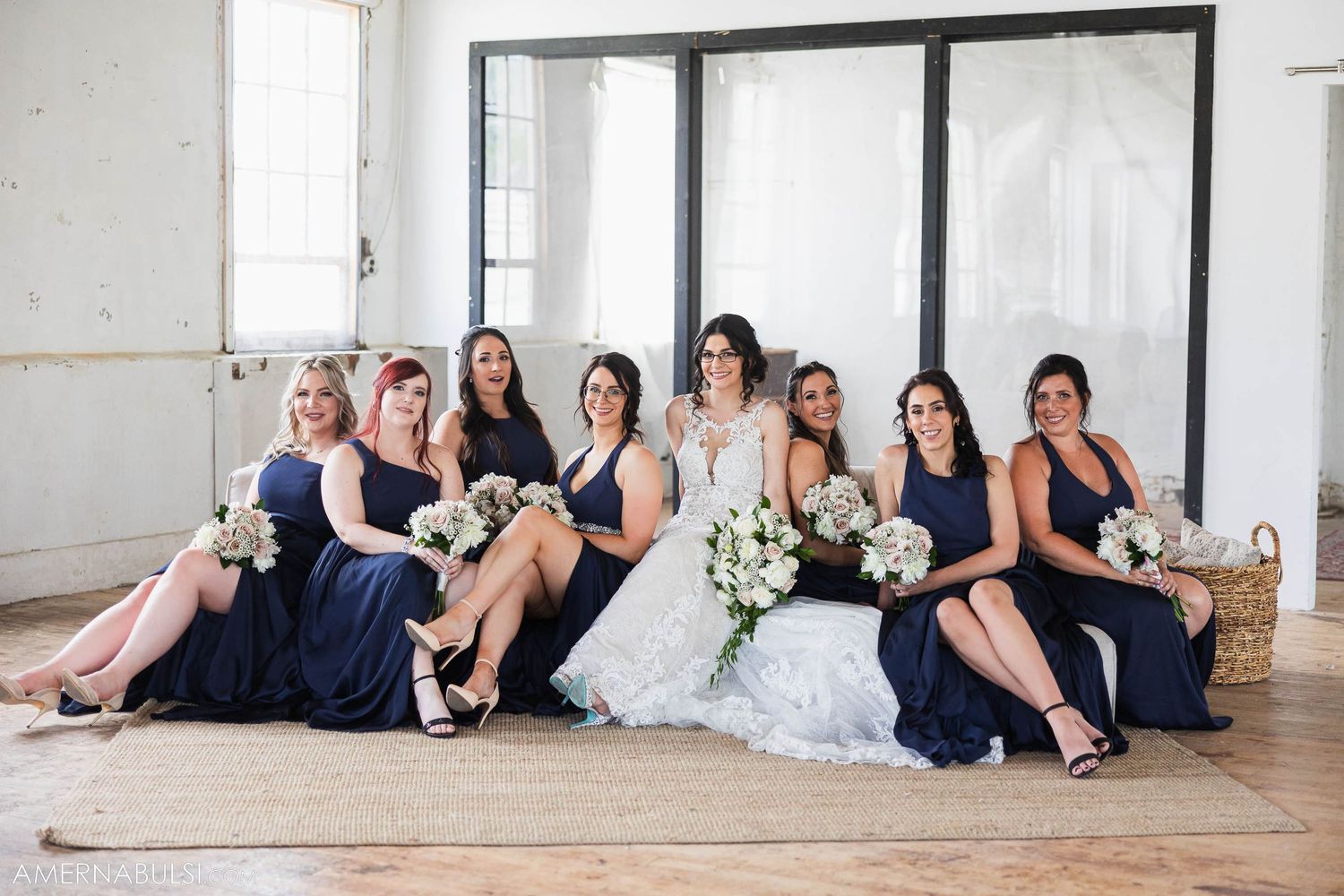 Bridesmaids in navy dresses relax with bride on elegant white couch.