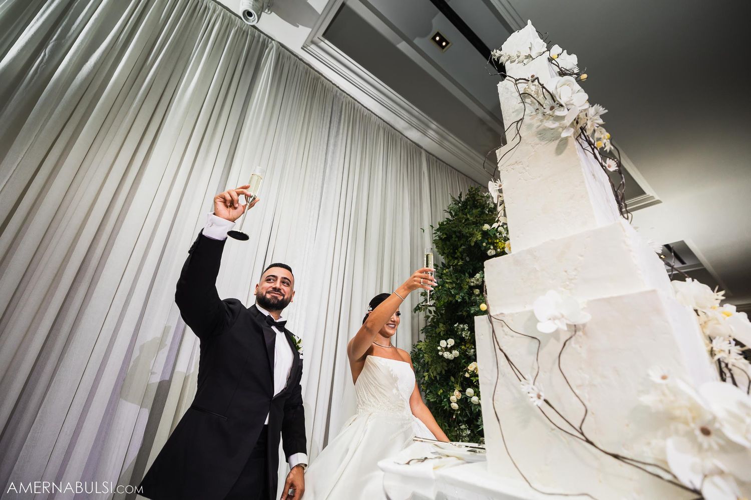Newlyweds celebrate next to their elegant white wedding cake with floral decorations.