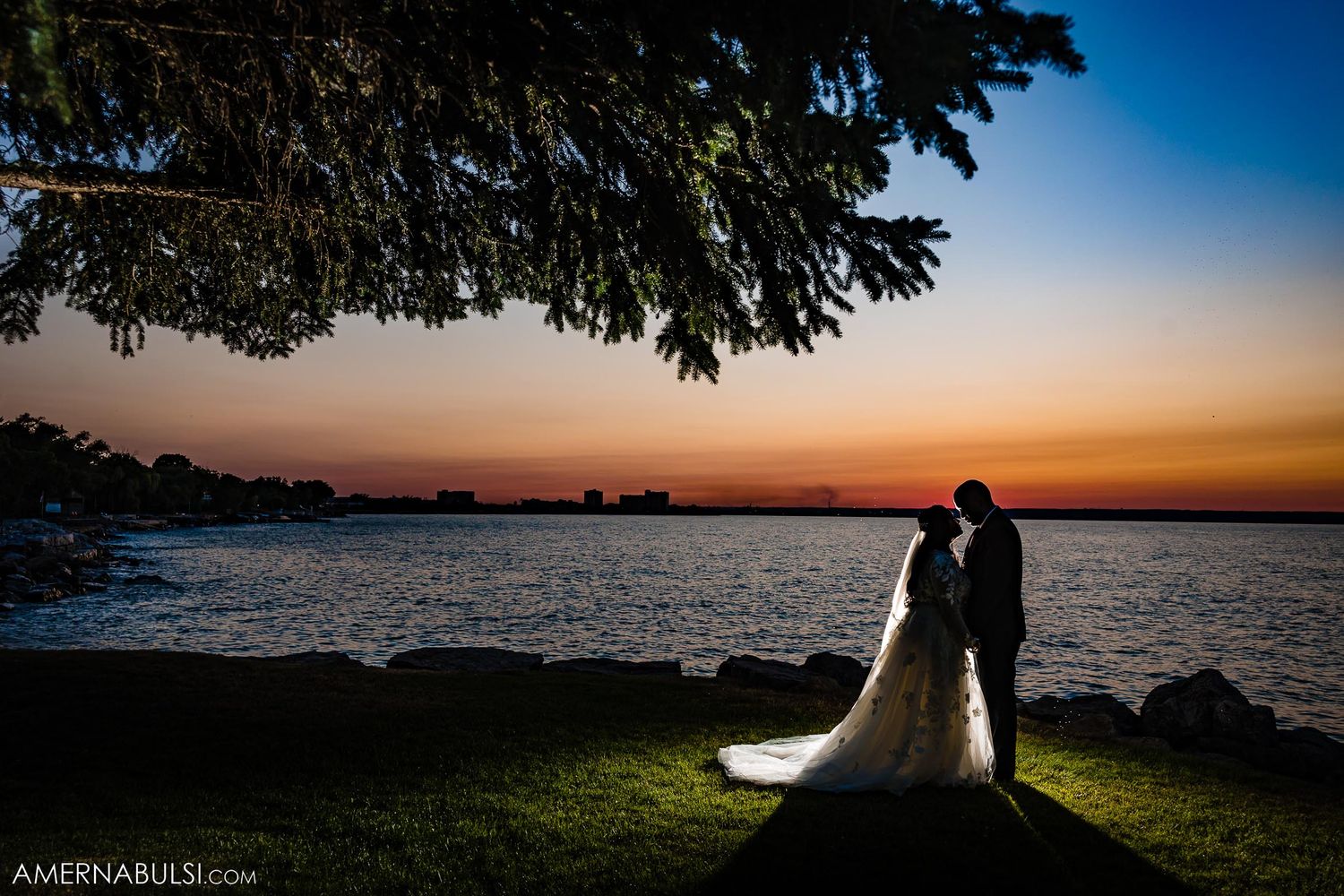 Silhouetted wedding couple standing by water during dramatic sunset.