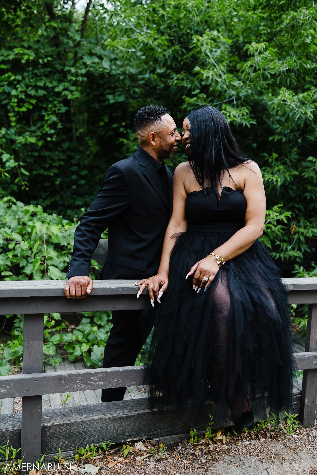 Romantic couple in black formal wear share an intimate moment on a wooden bench.