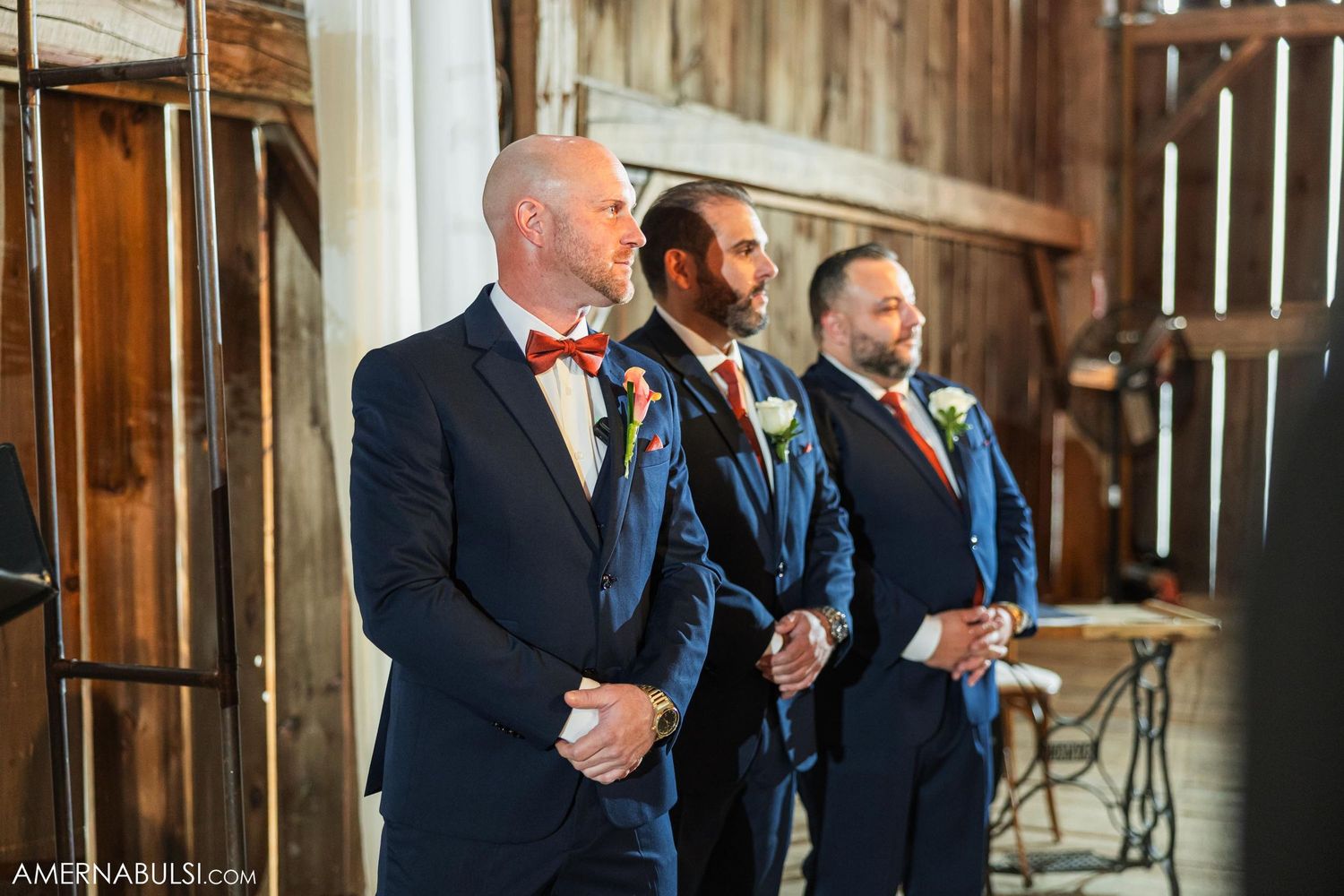 Groomsmen in formal suits and bow ties stand at attention during barn wedding ceremony.