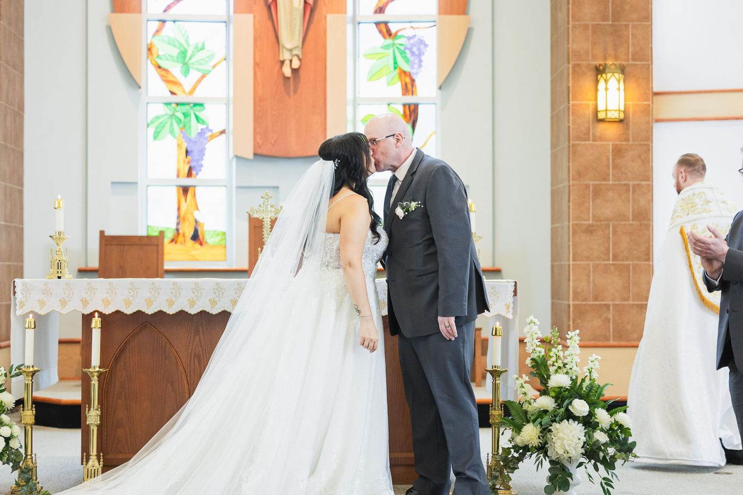 Wedding ceremony with stained glass windows and floral decorations in church setting.