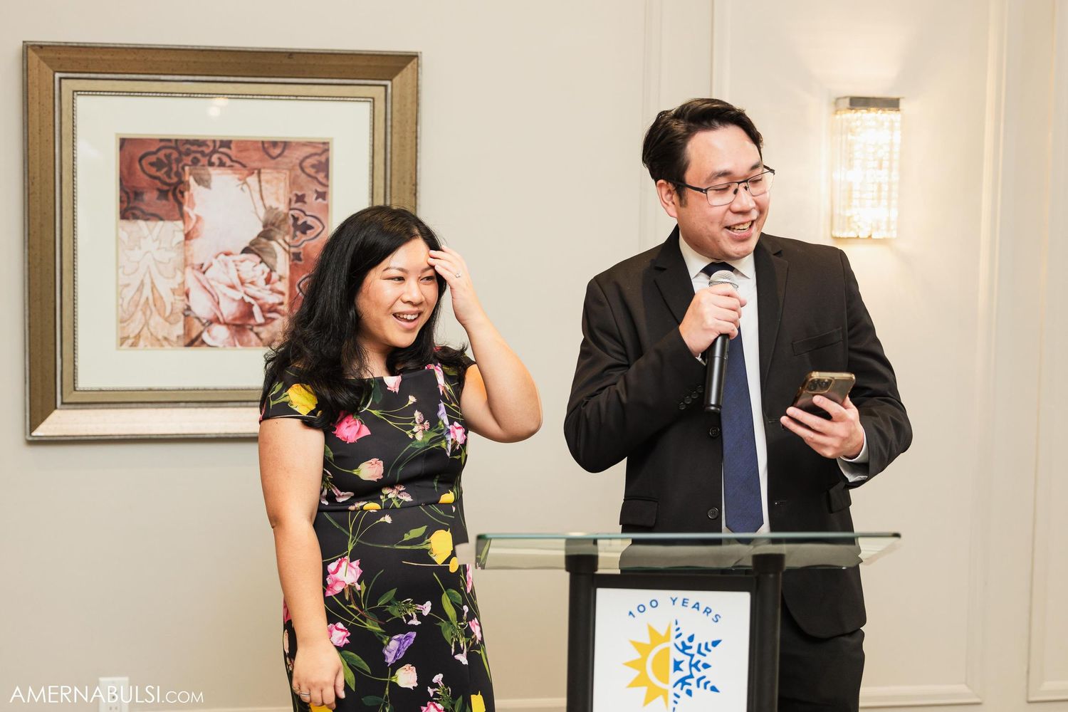 Speakers share a humorous moment at a wedding reception podium with decorative lighting in the background.
