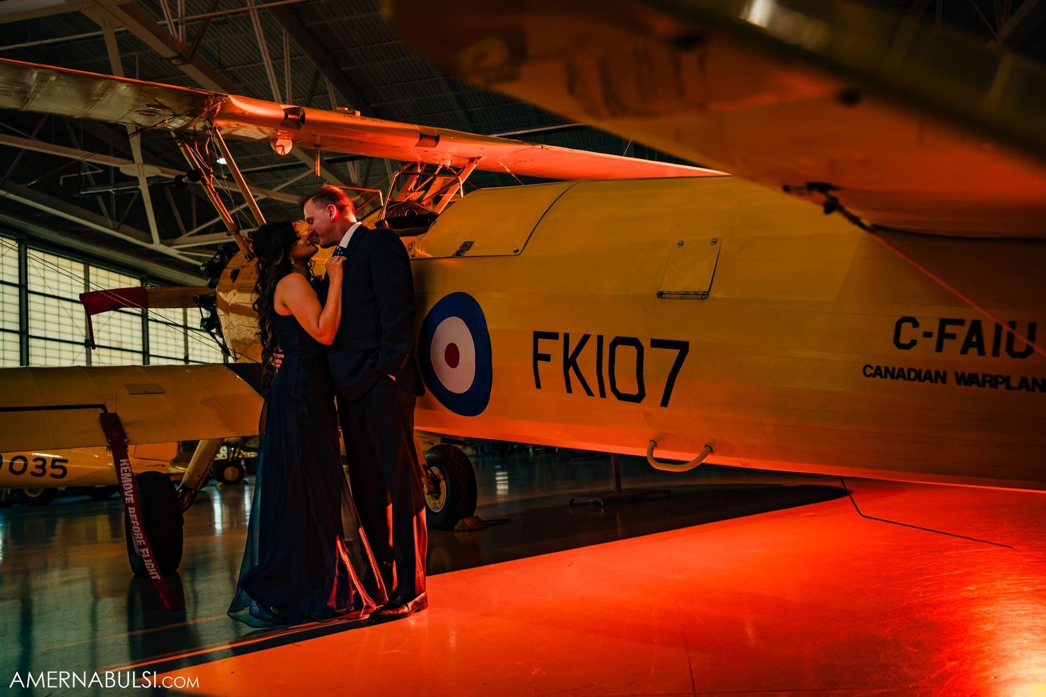 A couple embraces near a vintage aircraft under dramatic red lighting.
