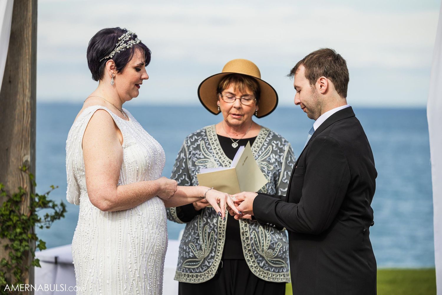 Outdoor wedding ceremony by the sea with officiant in tan sunhat reading from papers.