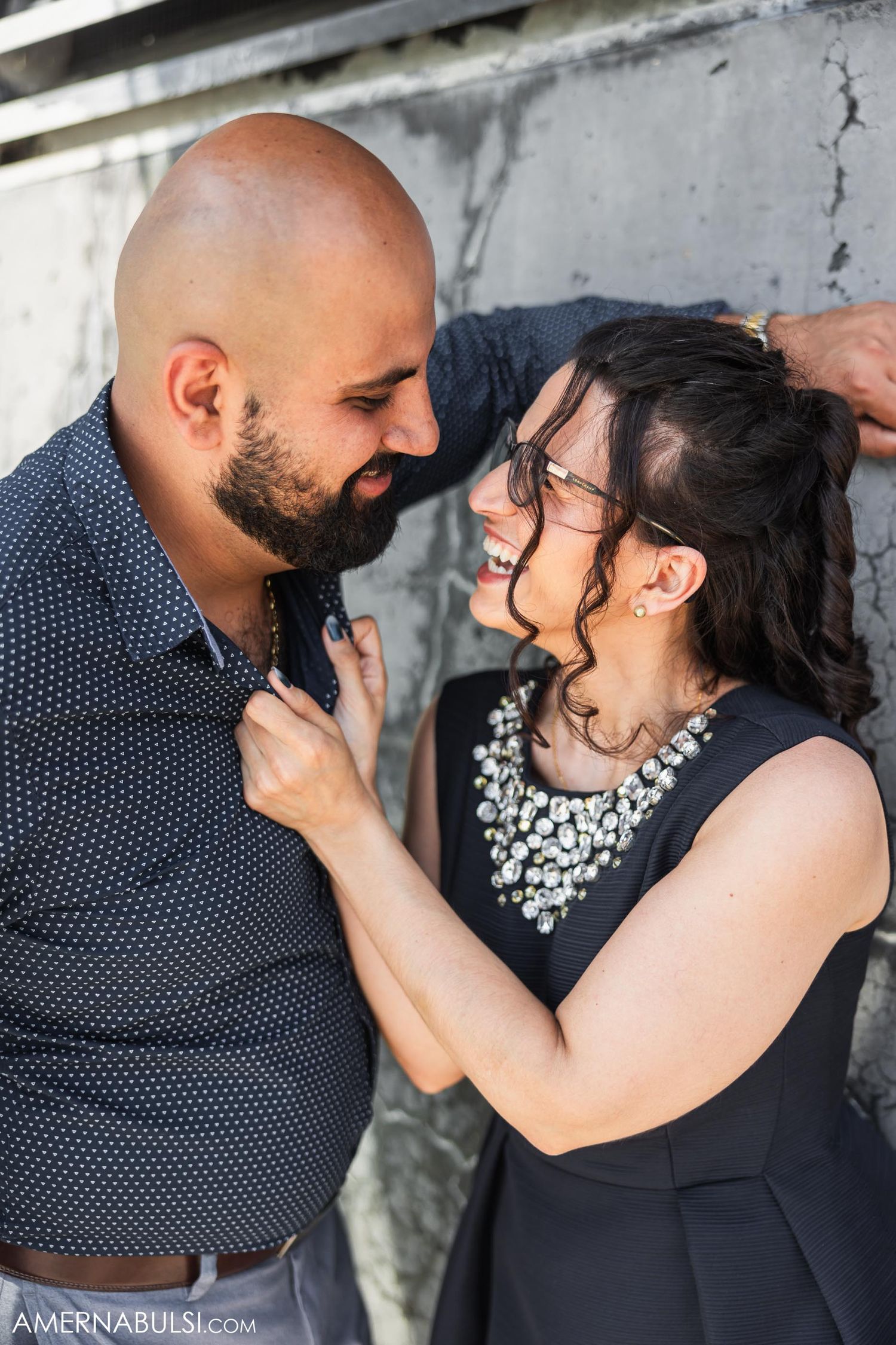 A couple shares a laugh during their engagement photo shoot against a concrete wall.
