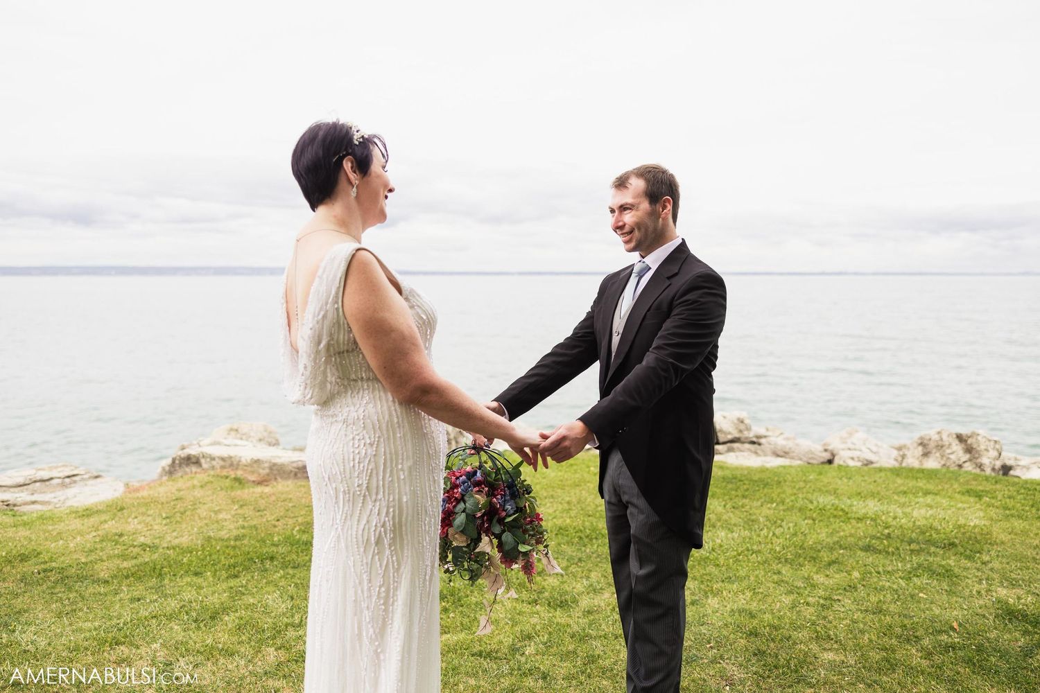 Couple shares intimate moment on grassy waterfront during wedding ceremony.