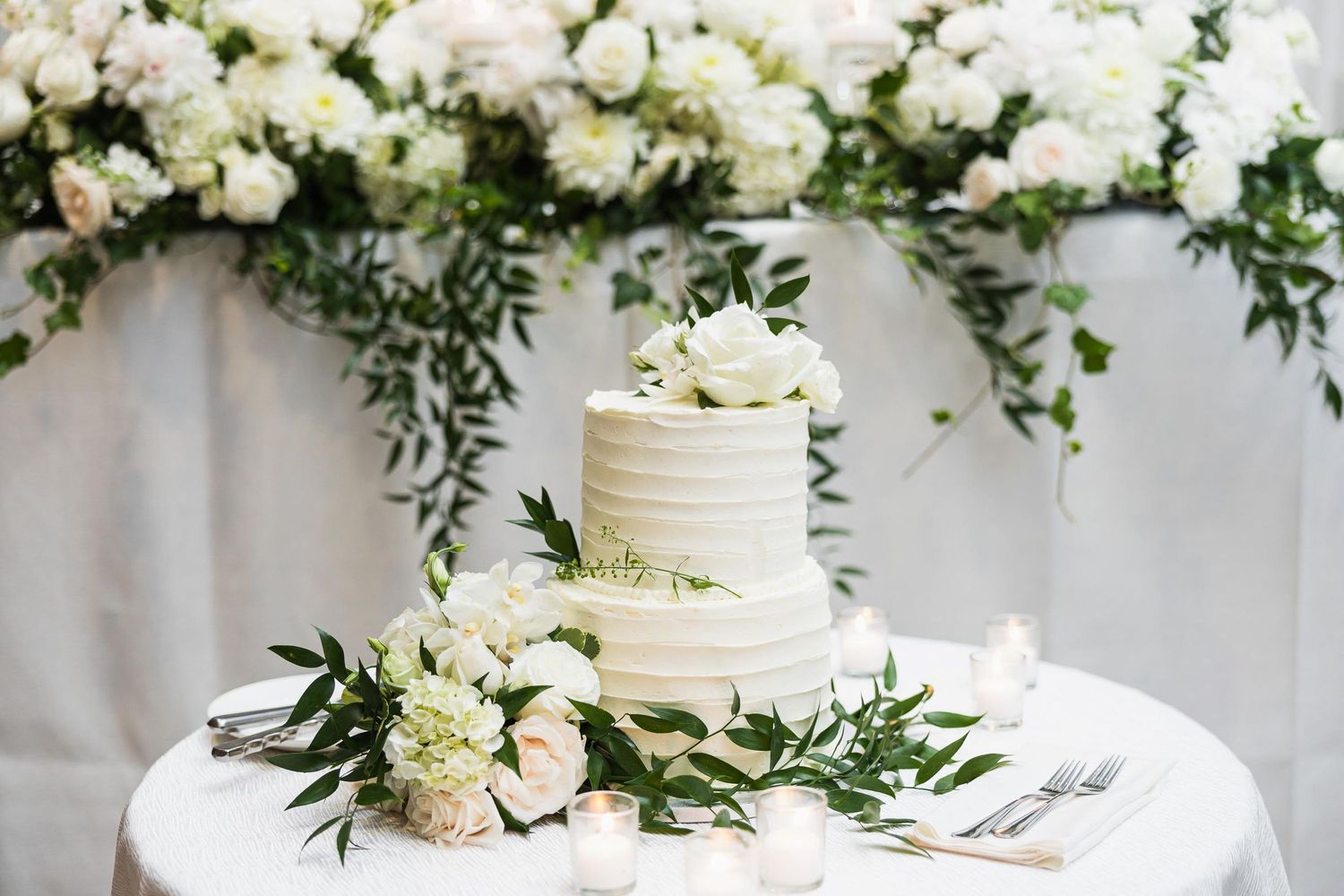 White buttercream wedding cake decorated with fresh flowers and greenery on round table.
