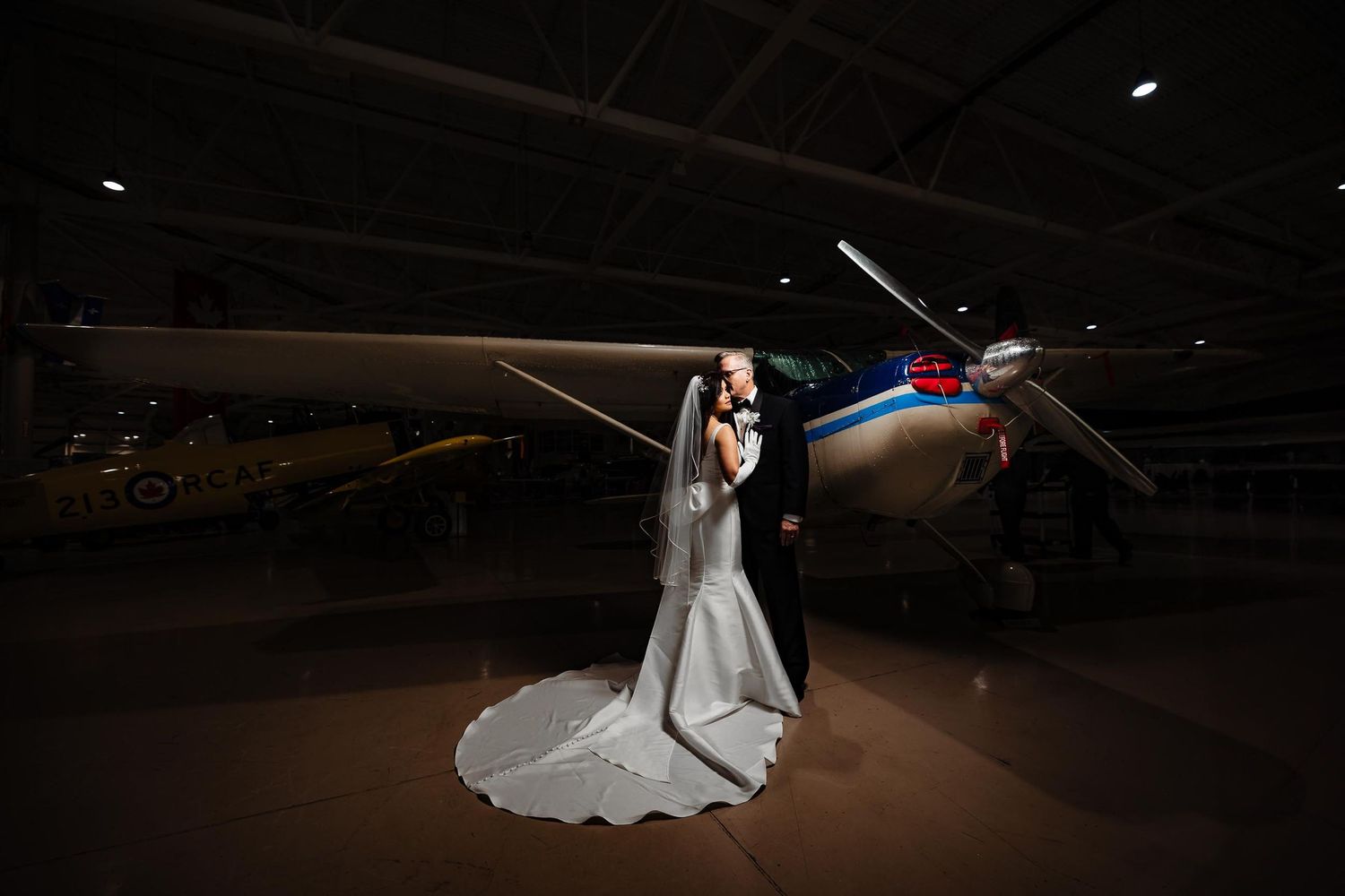 Wedding couple poses in front of vintage aircraft in dimly lit hangar creating dramatic silhouette.