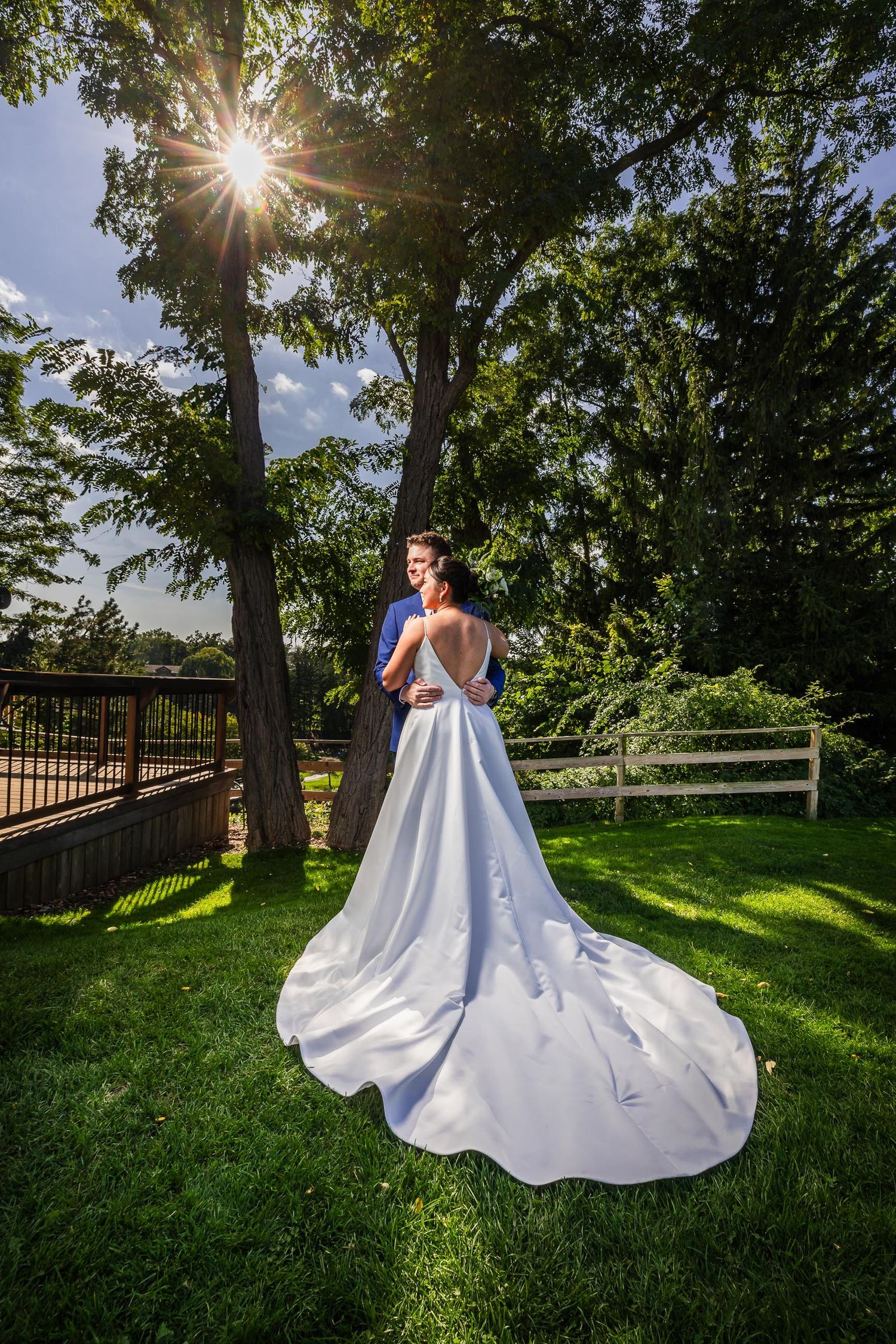 Bride in flowing white gown poses in garden with sunburst through trees.