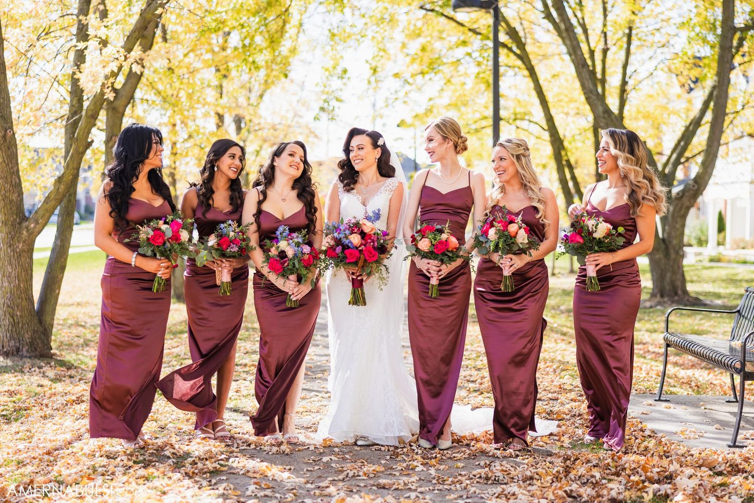 A bridal party in burgundy dresses poses together with bouquets during autumn.