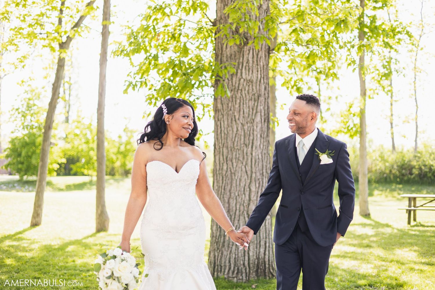 Elegant wedding couple walks hand in hand through a sun-lit wooded area.