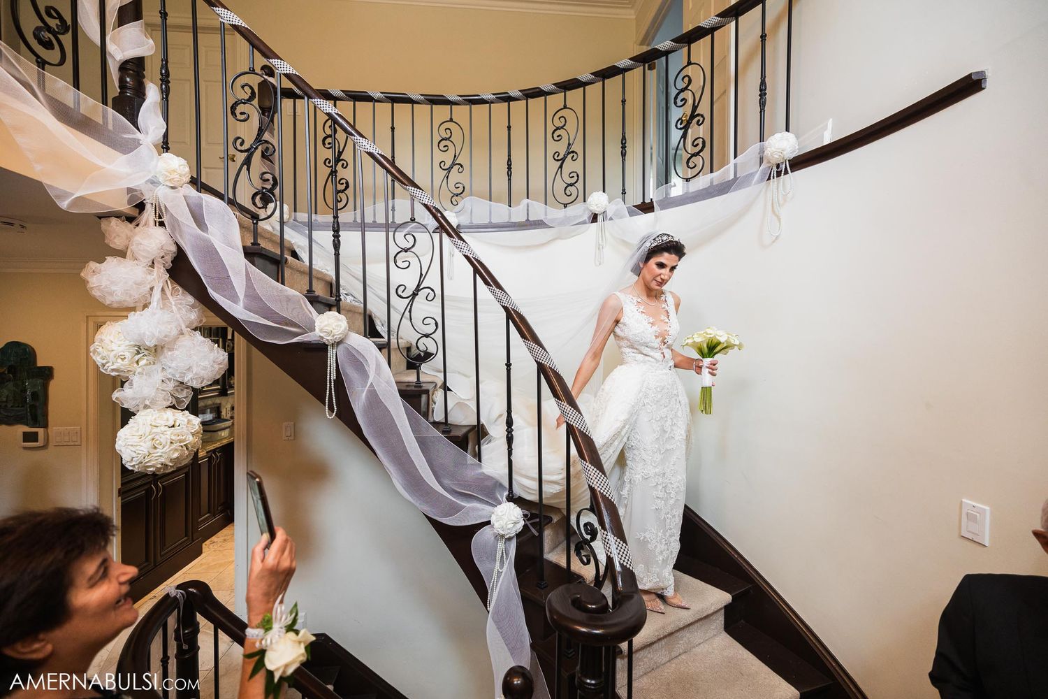 Bride descends elegant spiral staircase decorated with white flowers and fabric draping.