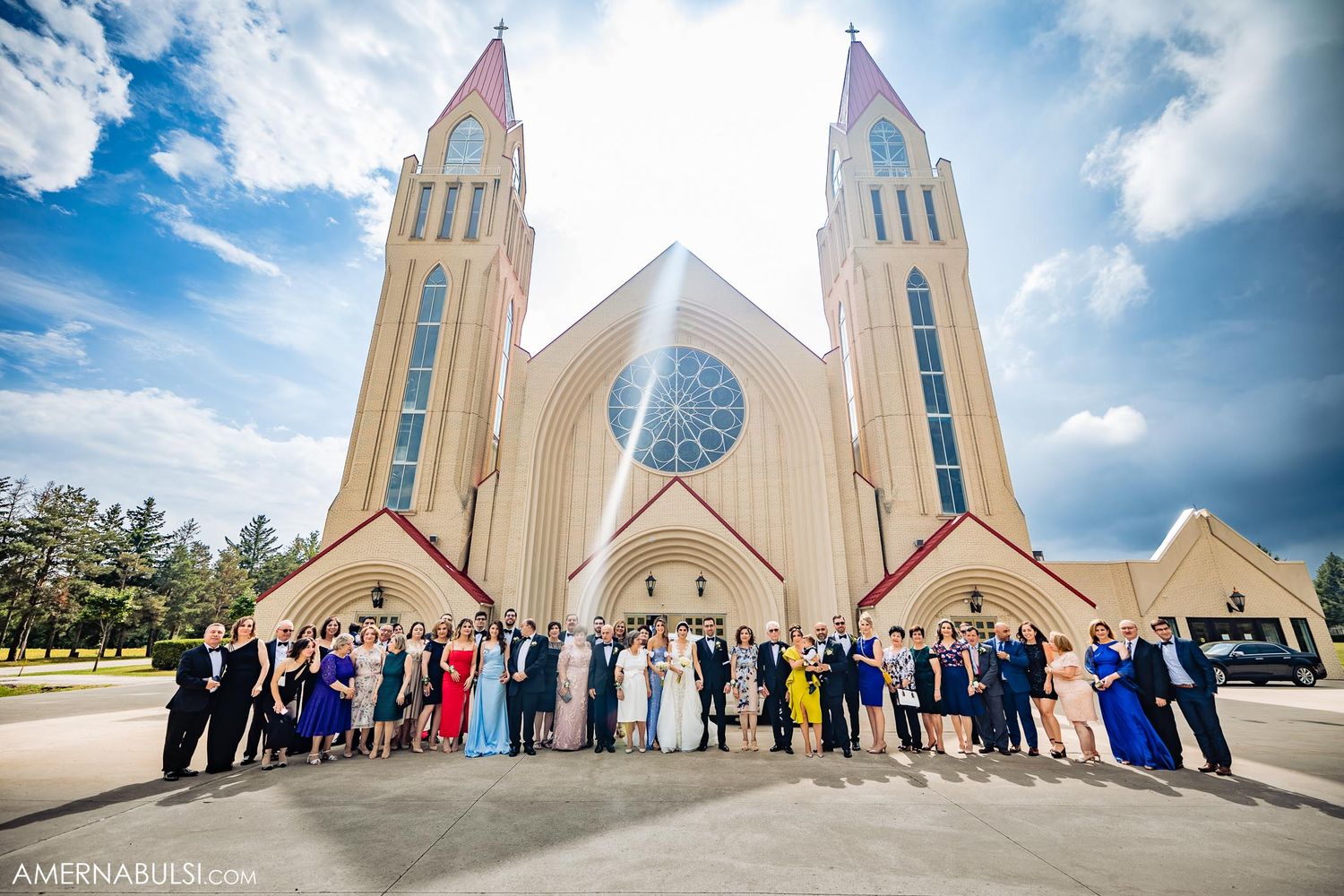 Large wedding party posed in front of modern cathedral with twin spires under blue sky.