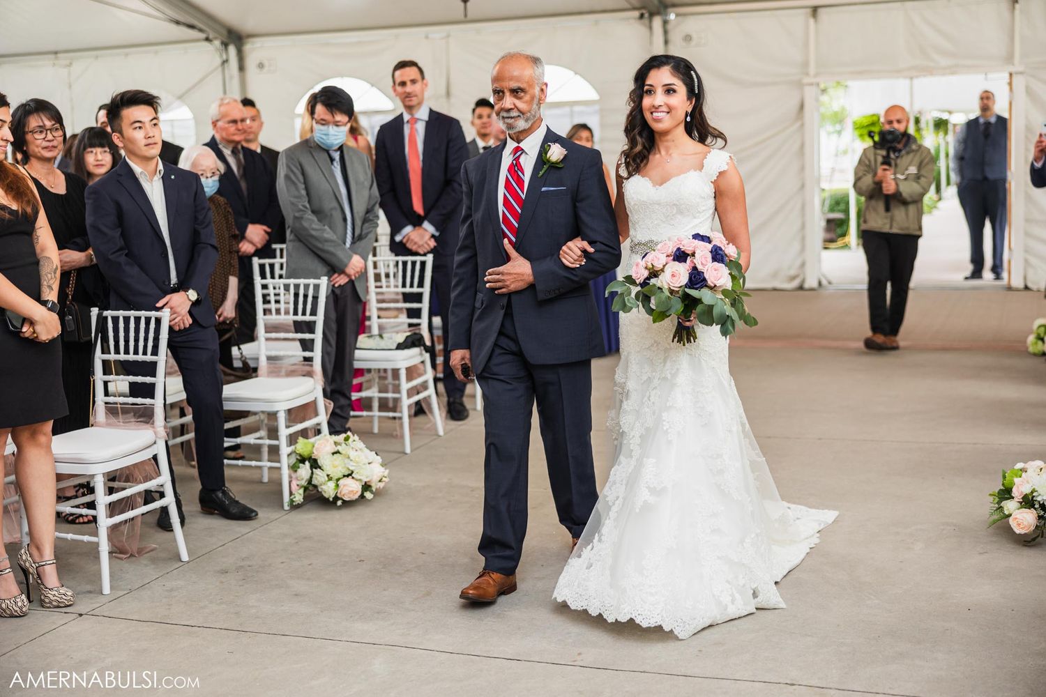 A wedding processional in a white tent with elegant floral arrangements and white chiavari chairs.