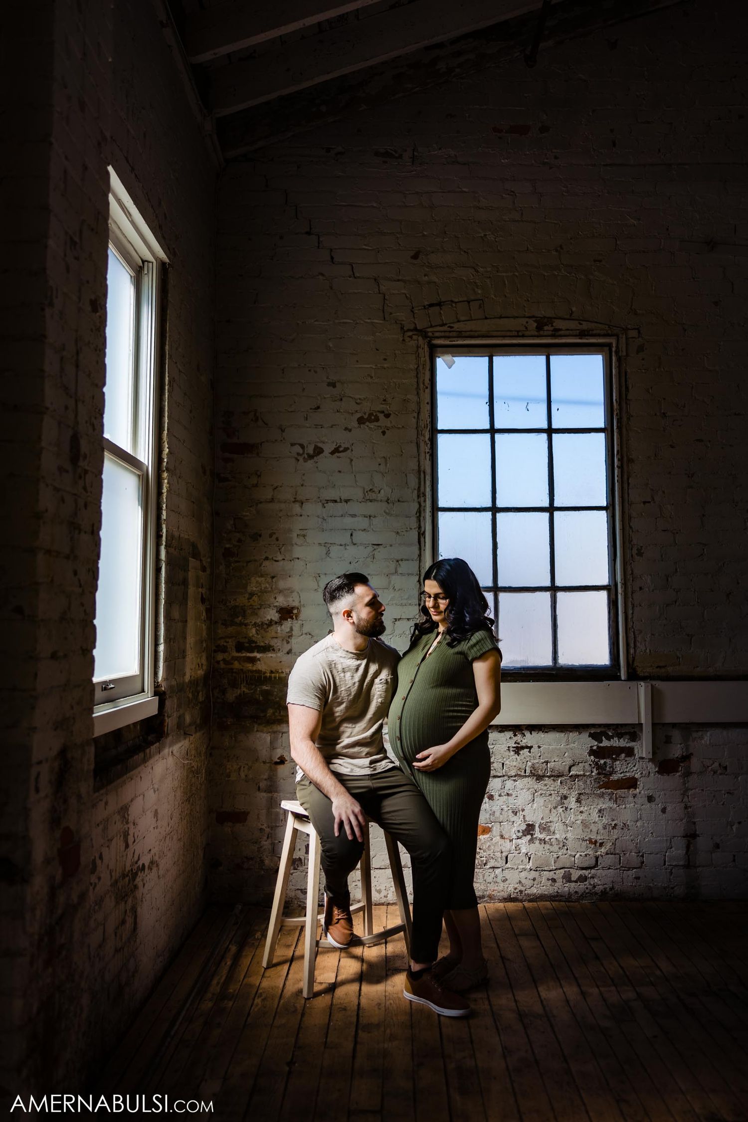 Dramatic maternity photo in rustic brick room with large windows and natural lighting.