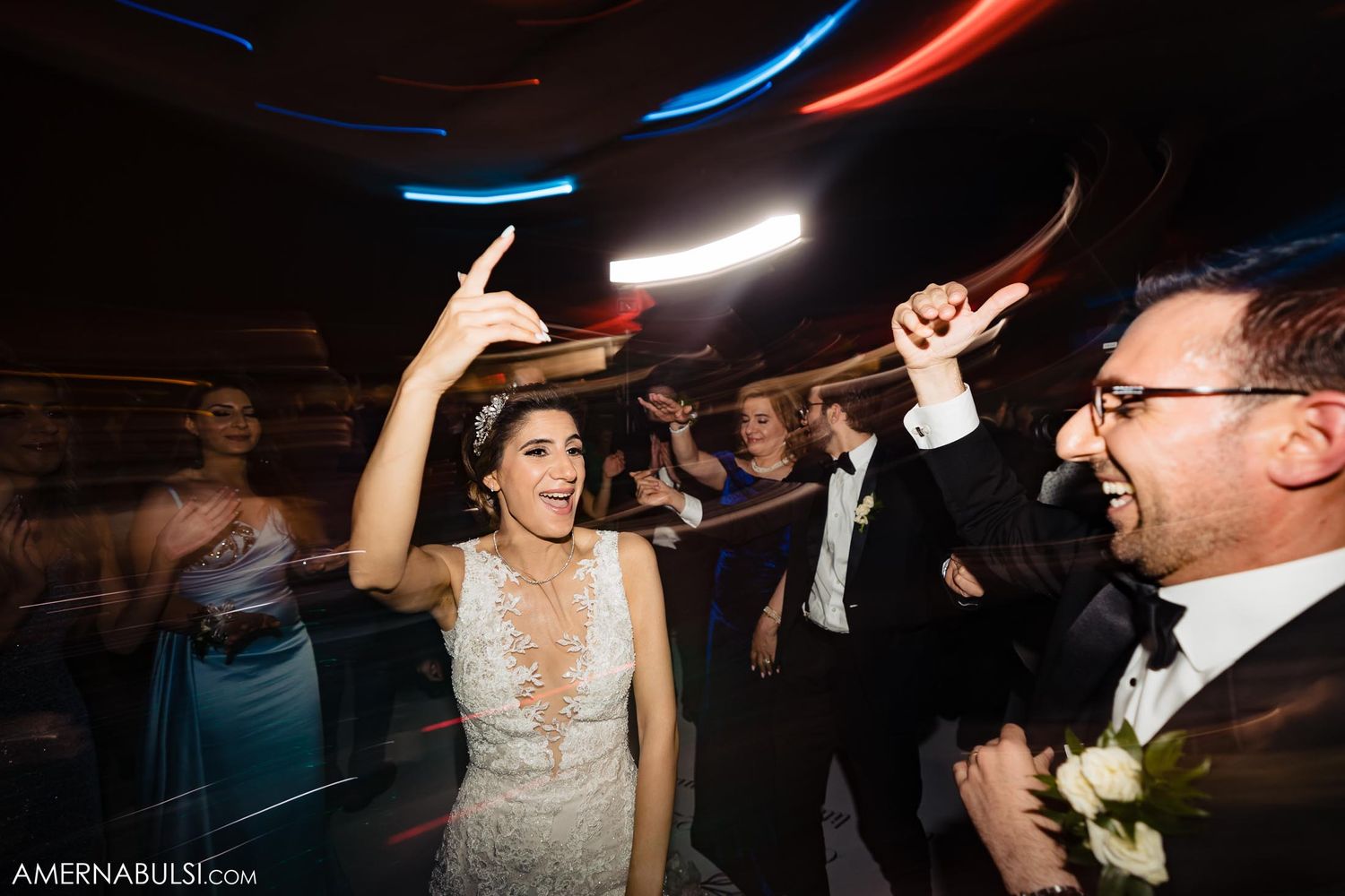 Energetic wedding reception dance party with bride and groom celebrating under colorful lights.
