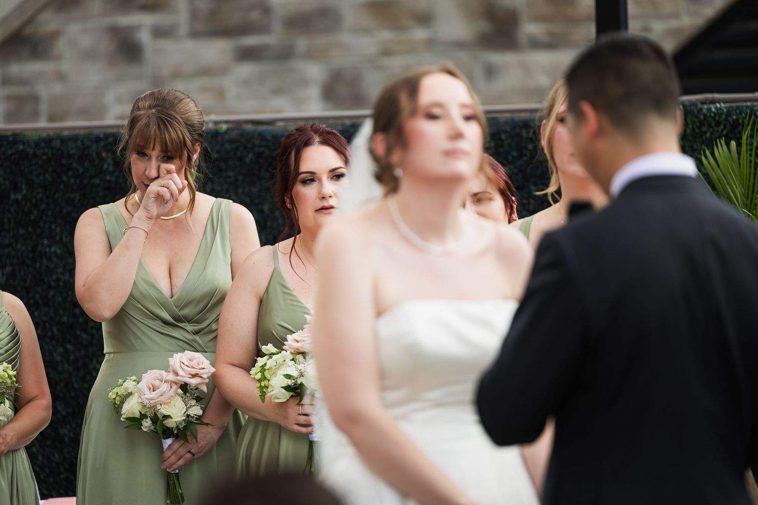 Emotional bridesmaids in sage green dresses watch wedding ceremony, one wiping tears, as bride and groom stand in foreground.