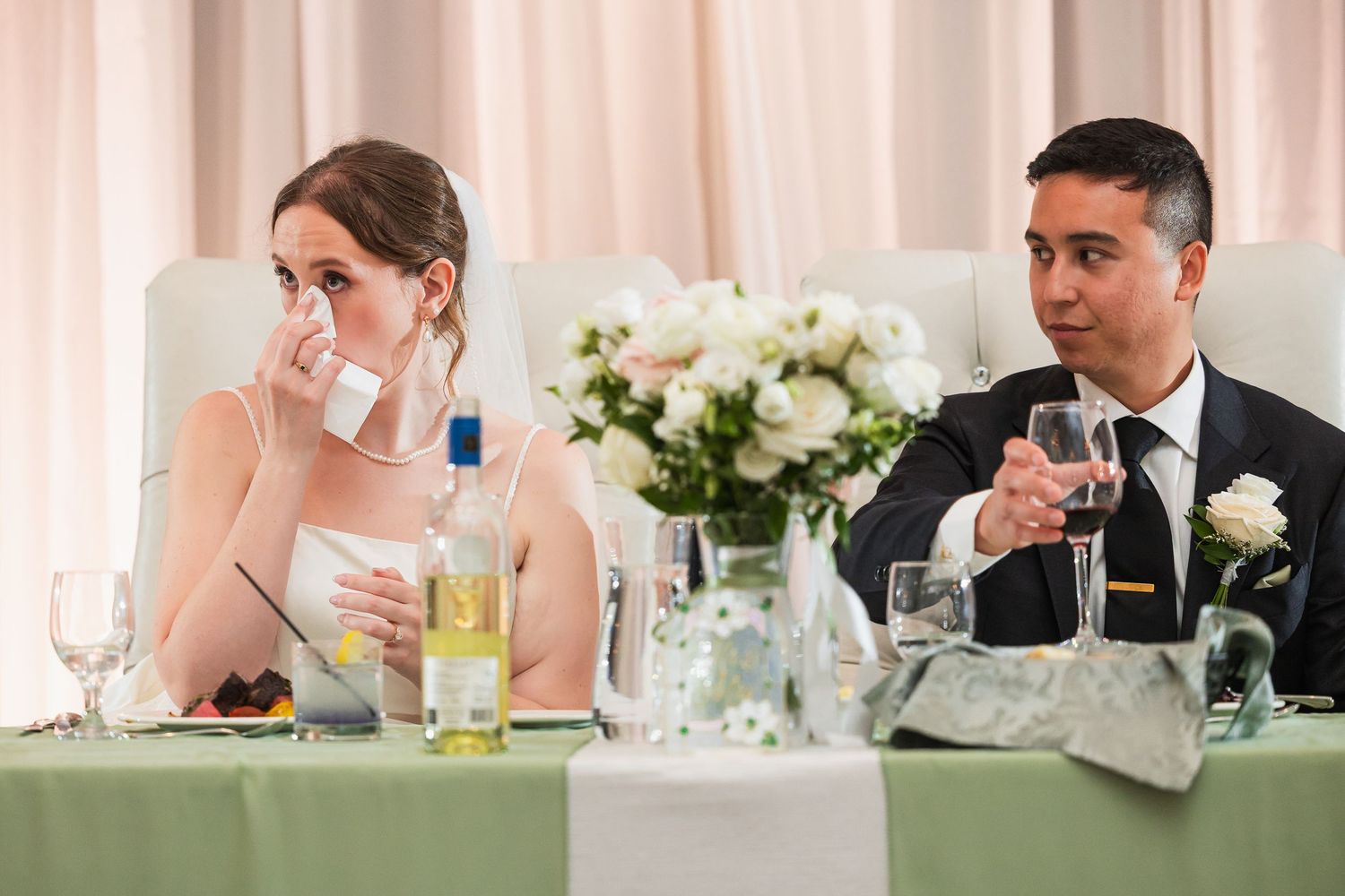Bride wiping tears while groom holds champagne glass at wedding reception head table with white floral centerpiece.