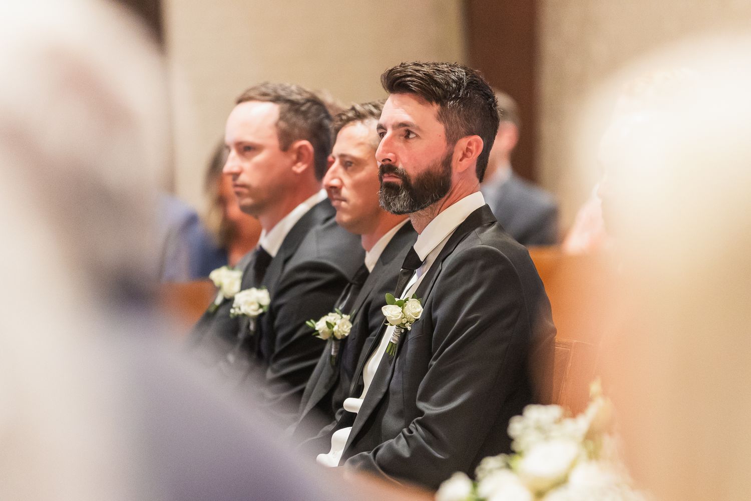 Groomsmen wearing boutonnieres sit attentively in church pews during wedding ceremony.