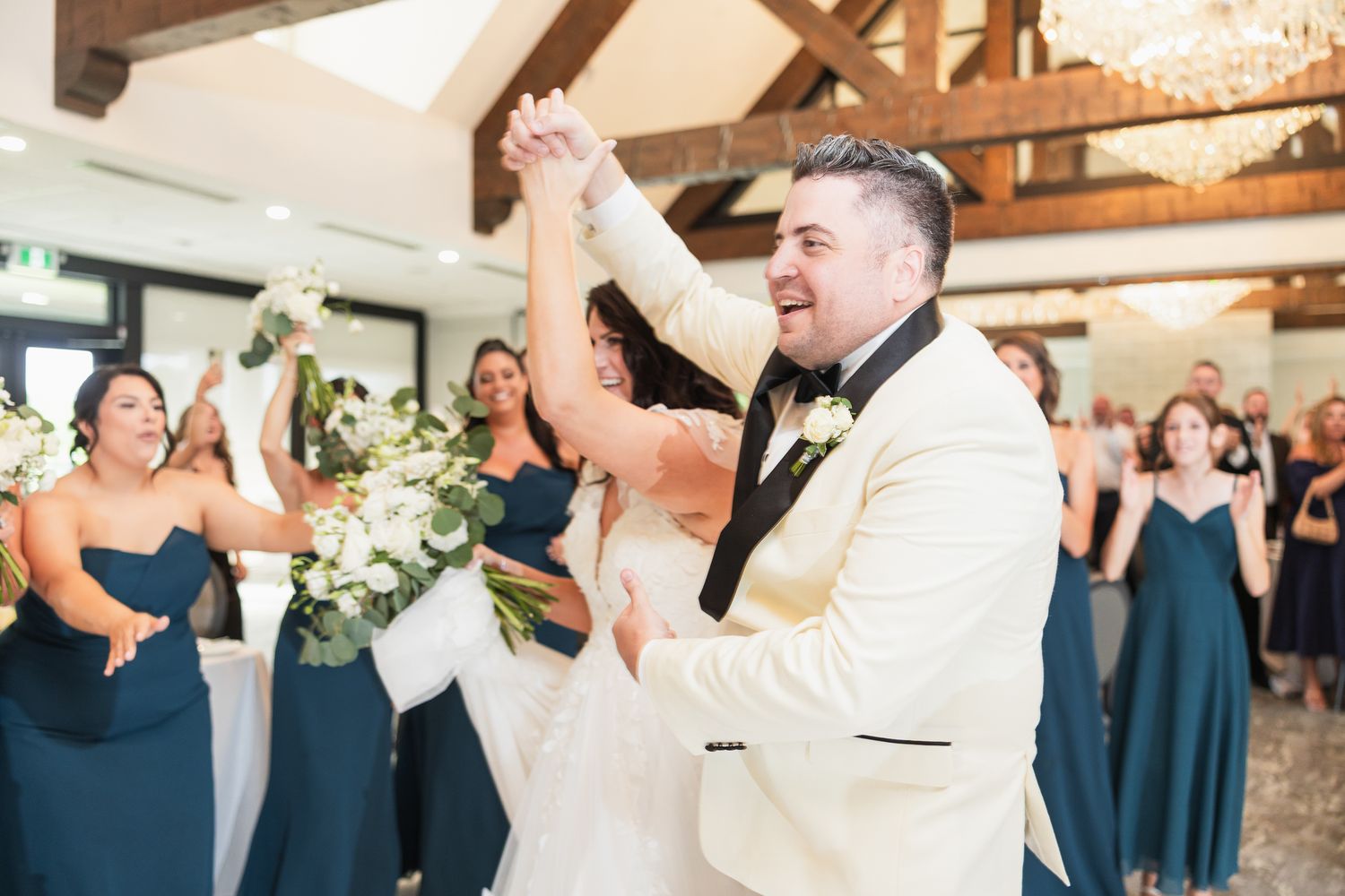 Joyful groom raises bride's hand in celebration surrounded by bridesmaids in teal dresses at wedding reception.