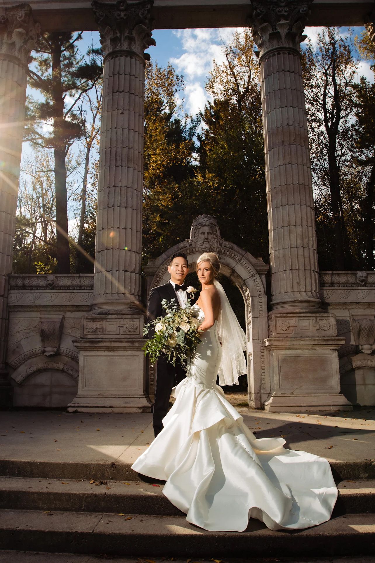 Wedding couple poses elegantly among classical stone columns during golden hour.