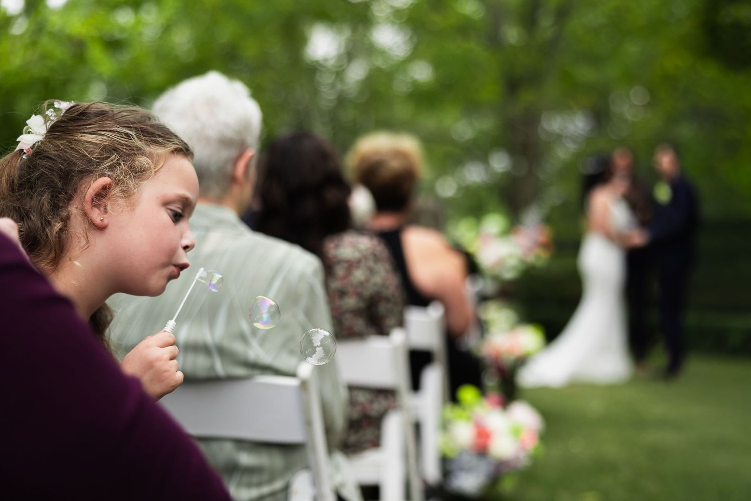 Young flower girl blowing bubbles during outdoor wedding ceremony while guests watch couple exchange vows.