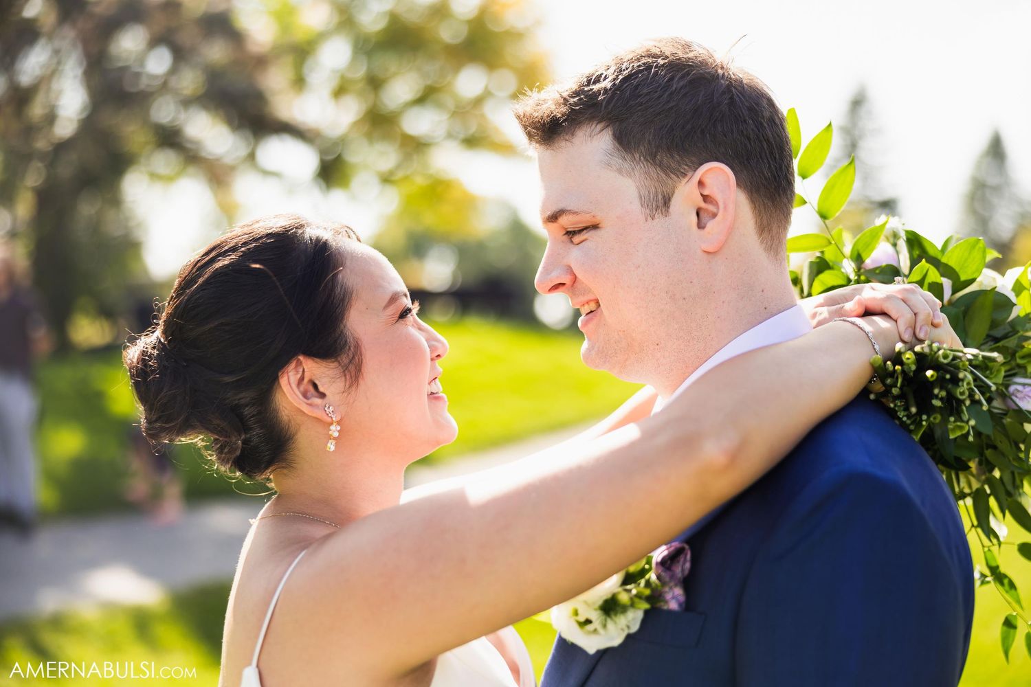 A couple embraces outdoors in bright sunlight with gardens in the background.