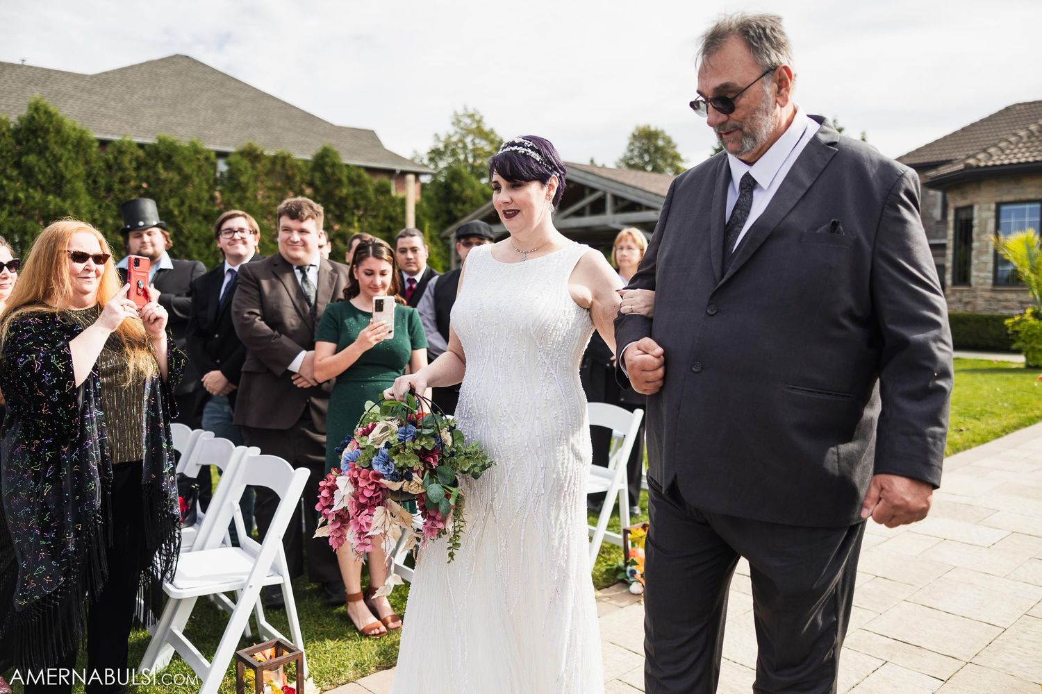 Wedding processional on stone pathway with white chairs and guests.