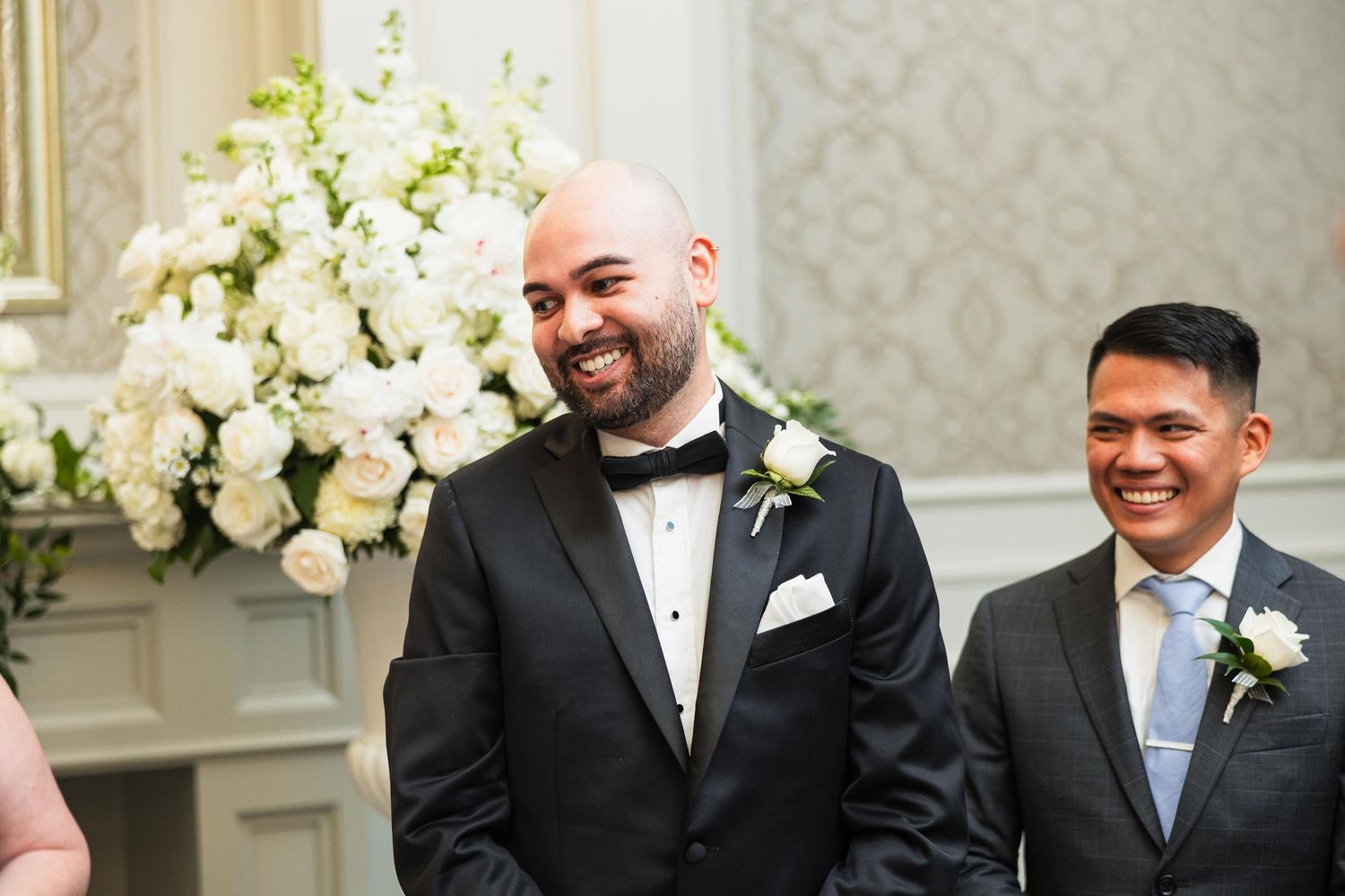Wedding party members share joyful moment by white floral arrangement in elegant venue.