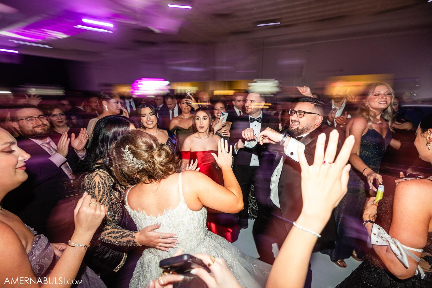 Crowded wedding reception dance floor with guests celebrating under purple venue lighting.