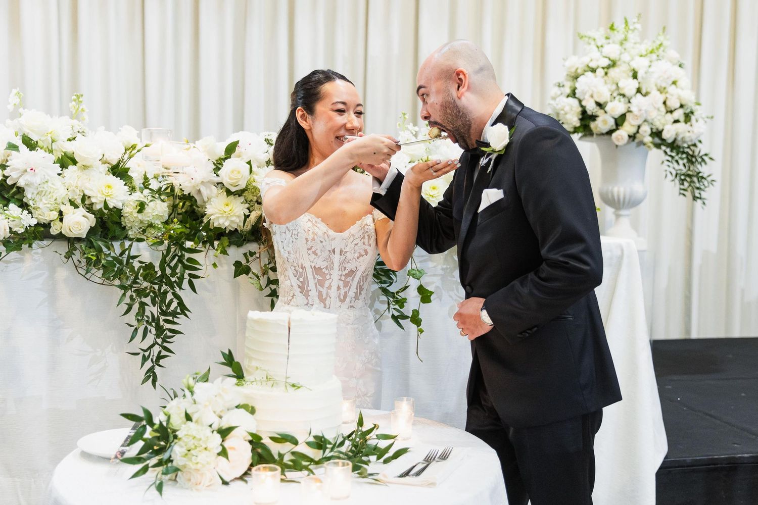 Newlyweds share cake-cutting moment at reception with white floral decorations.