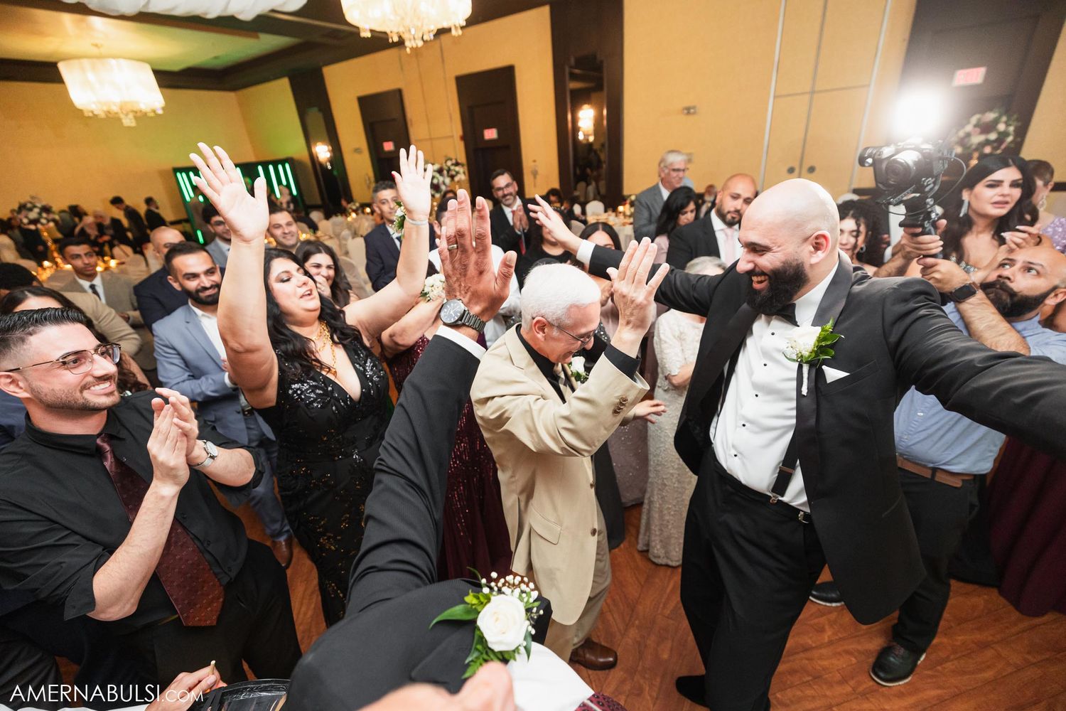 Energetic wedding guests and a couple dancing enthusiastically at a reception with warm lighting and decorative chandeliers.