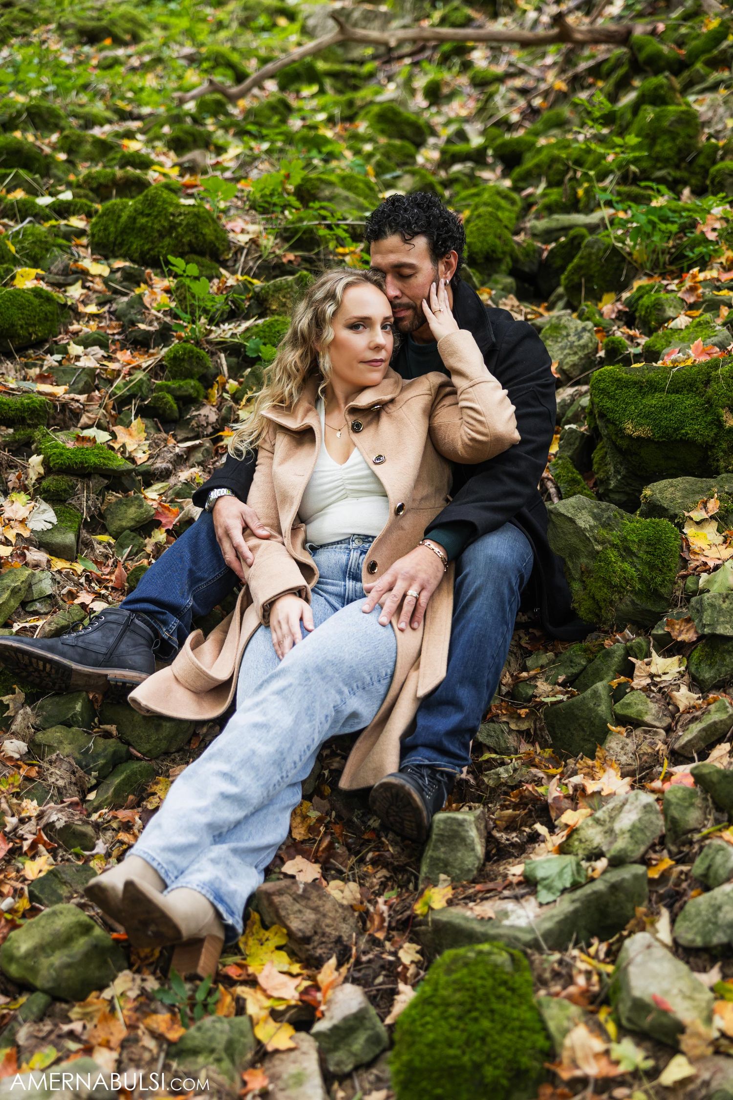 Couple in casual autumn wear sitting intimately among mossy rocks and fallen leaves.