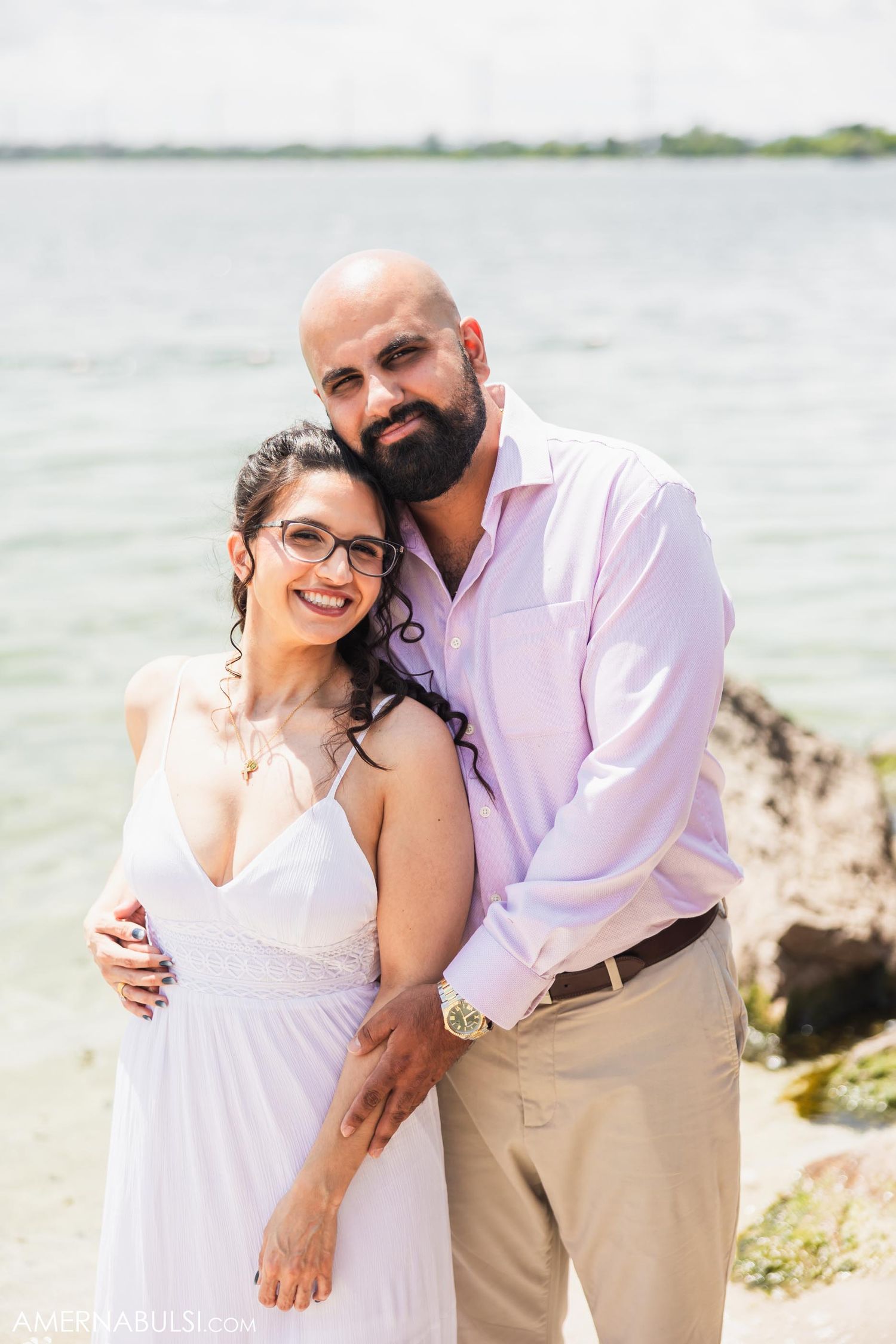 Happy couple pose together on rocky beach with water in background.