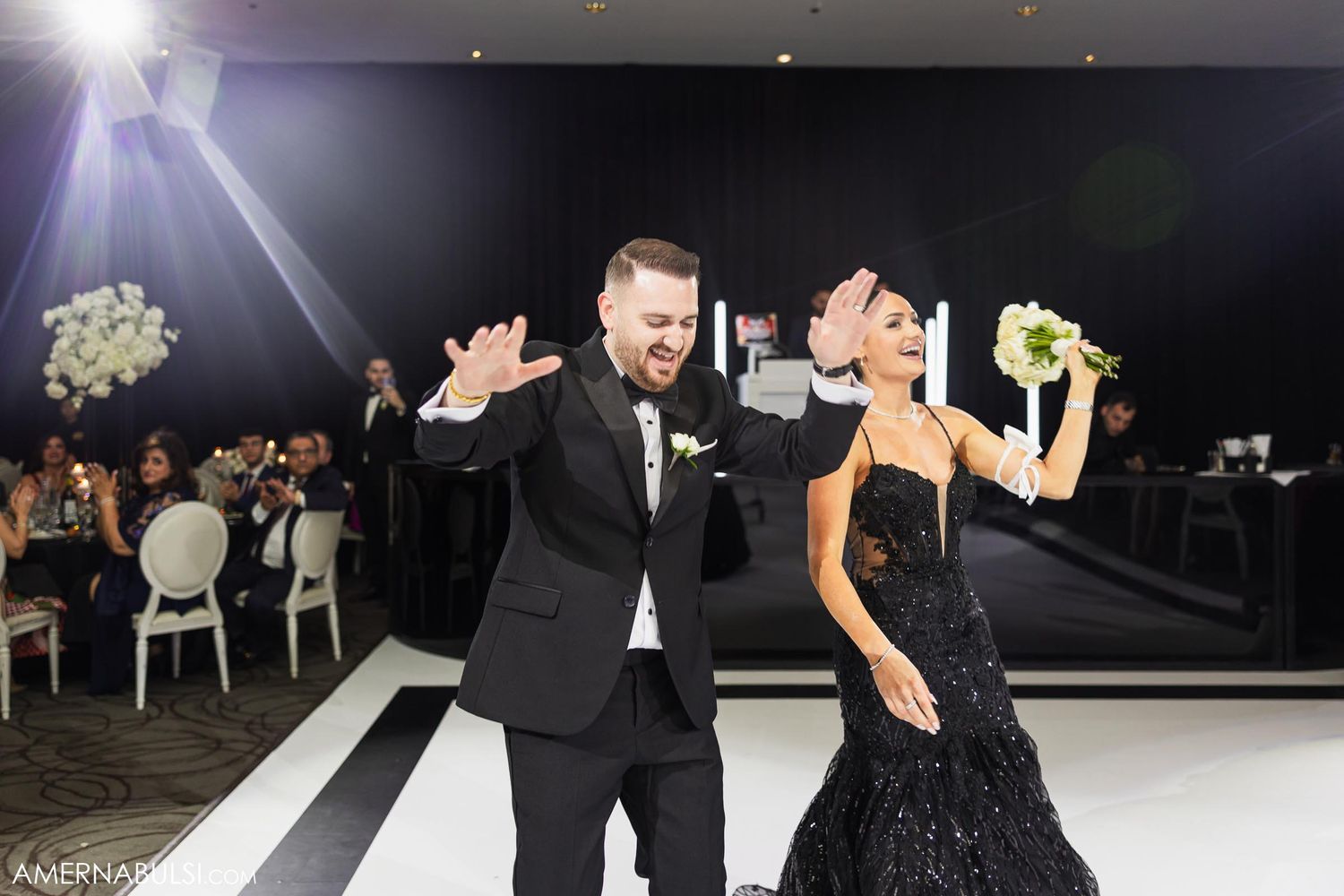 A couple in formal black attire celebrates energetically on a white dance floor at their reception.