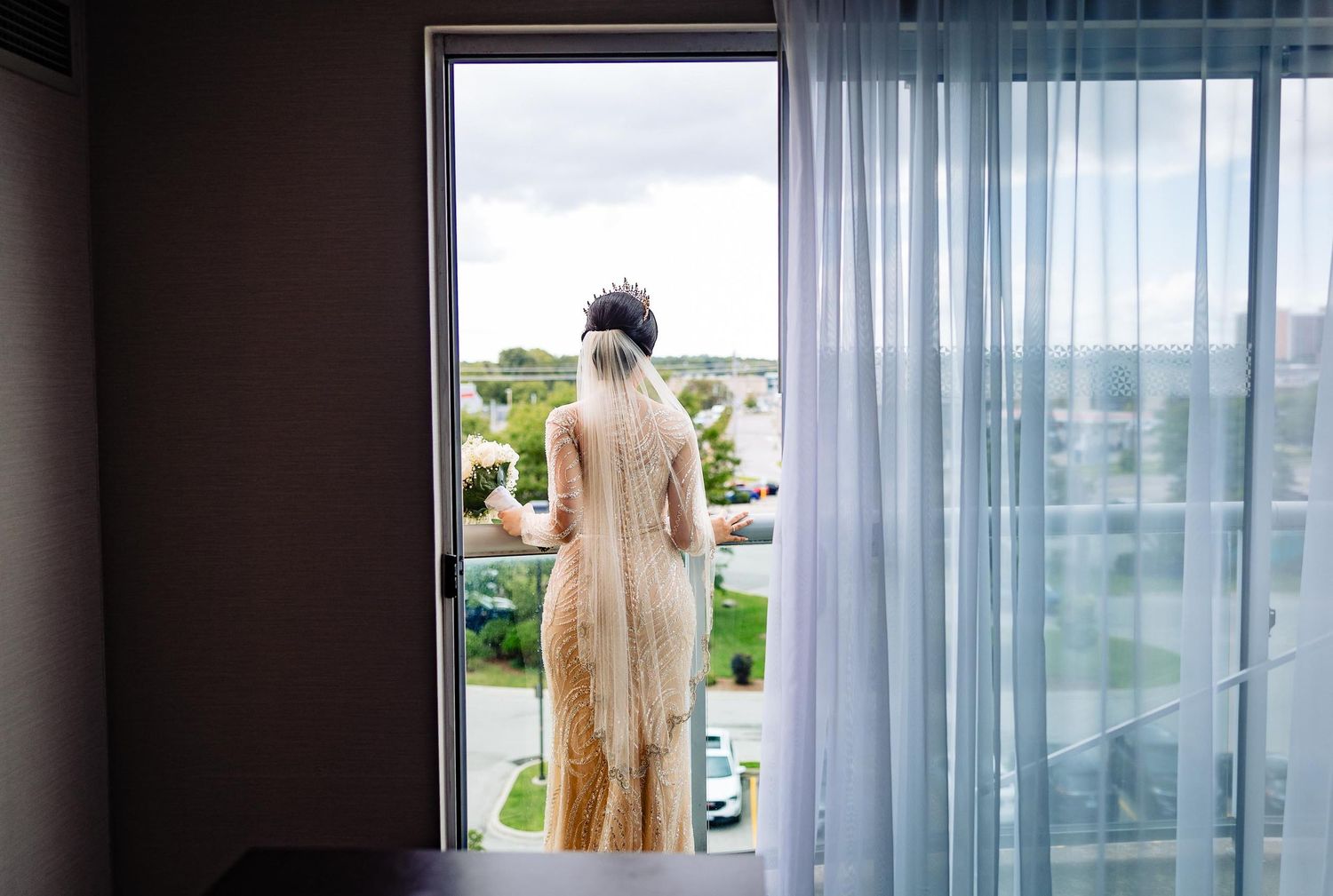 A figure in a cream-colored dress stands at a balcony doorway looking out at a sunny landscape.