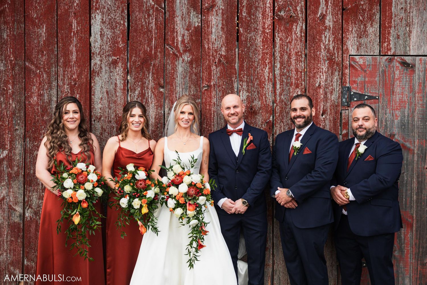 Wedding party lined up against rustic red barn wearing navy and burgundy attire.