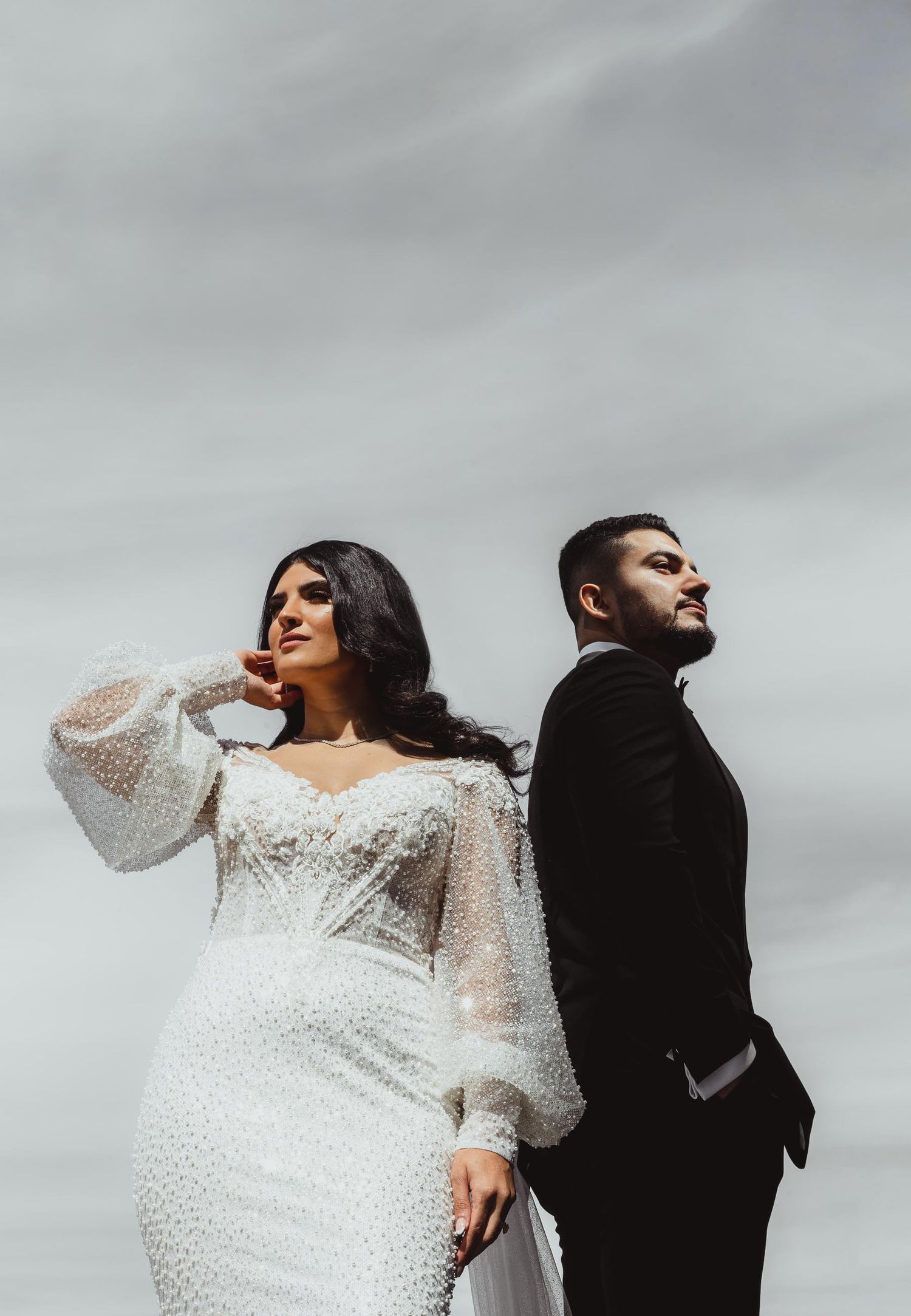 Stylish bride in beaded gown and groom in black suit pose back-to-back against cloudy sky.