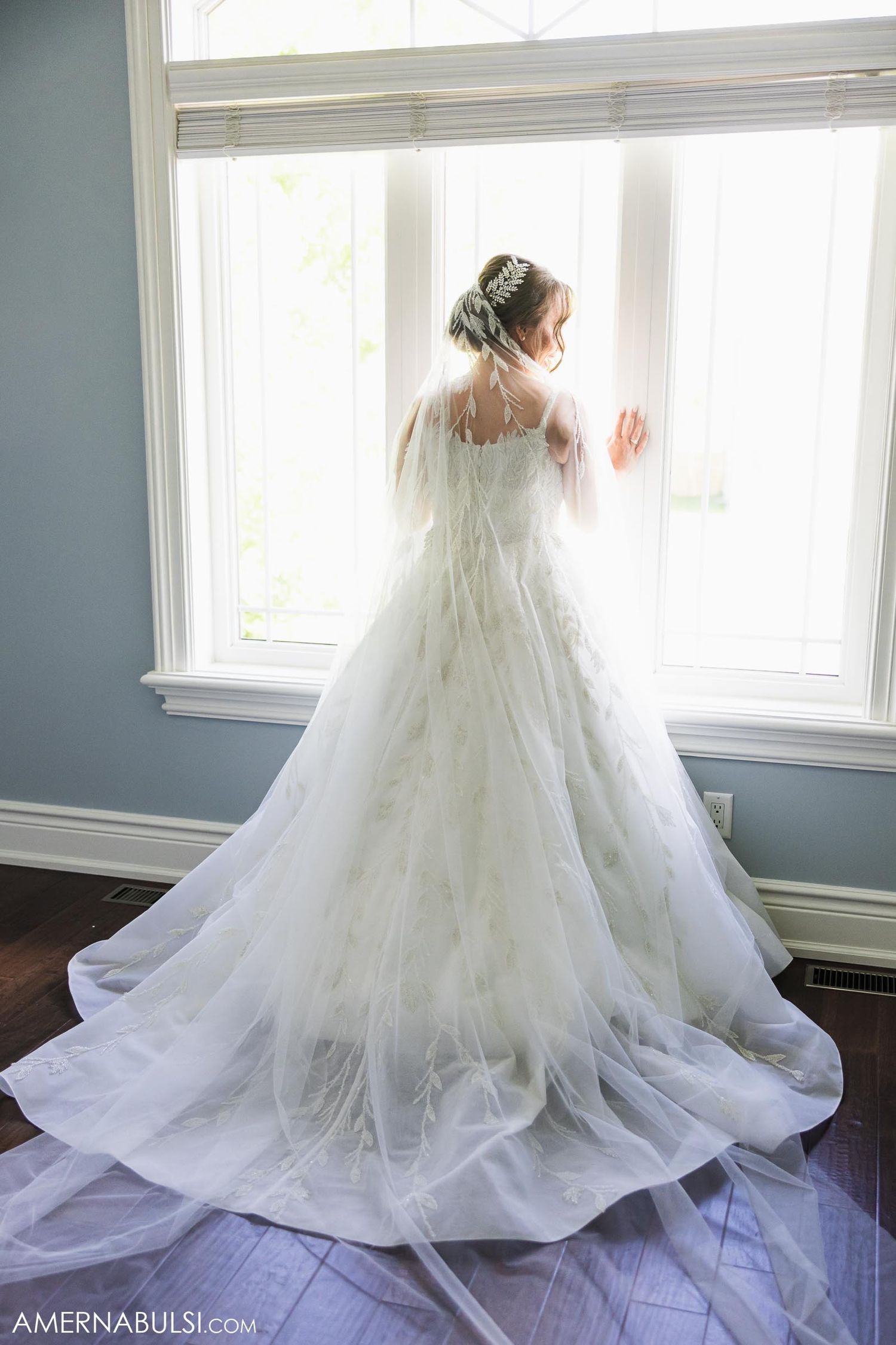 A flowing white wedding dress with delicate lace details captured against bright windows.