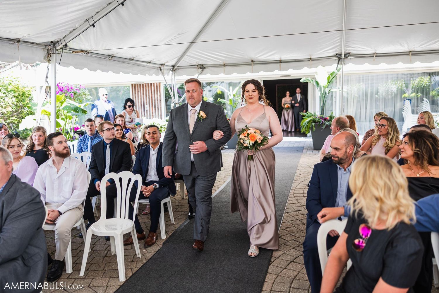 Wedding processional in a white tent with guests seated on white chairs along the aisle.