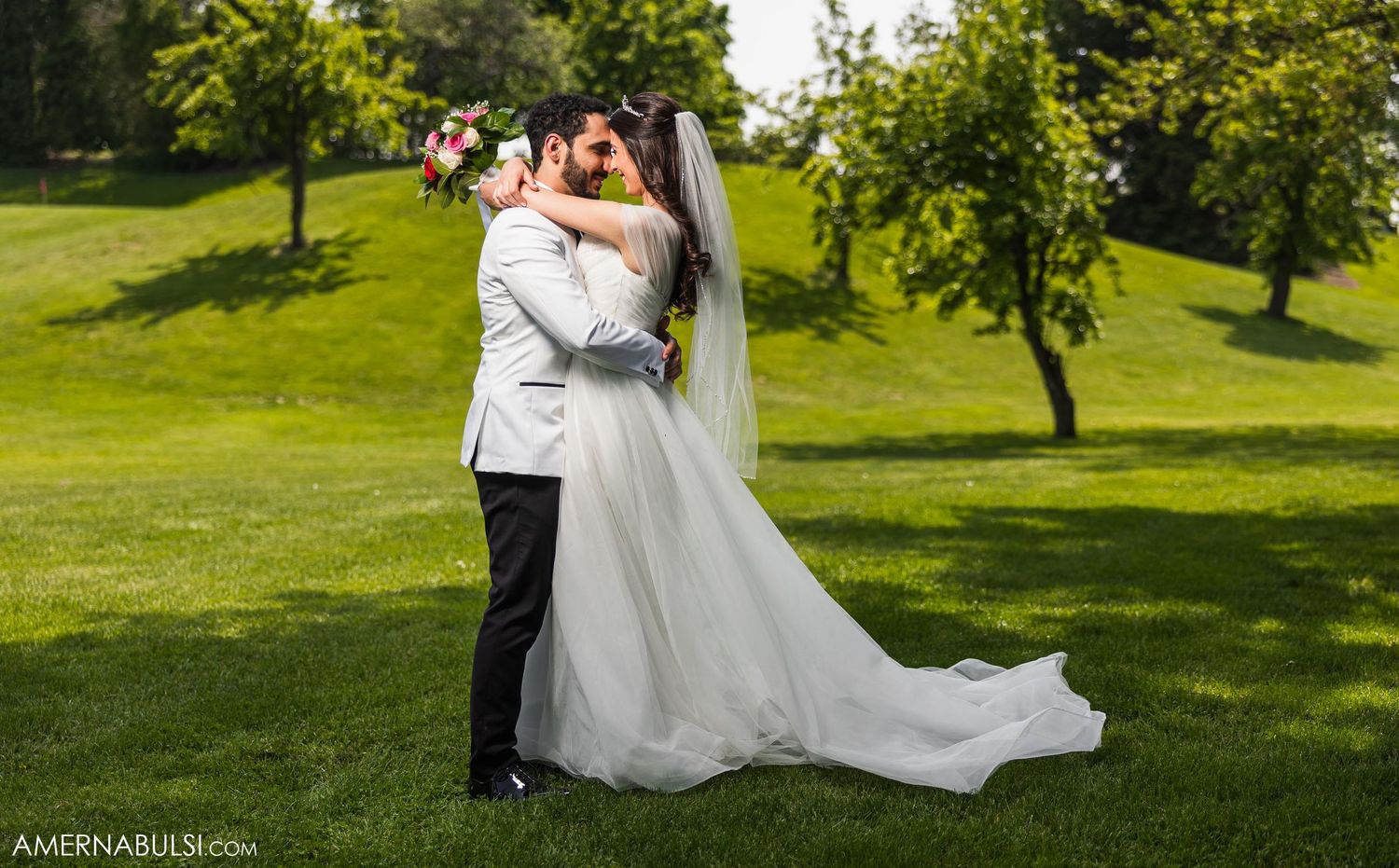 Wedding couple embracing in a romantic pose on a grassy hill surrounded by trees.