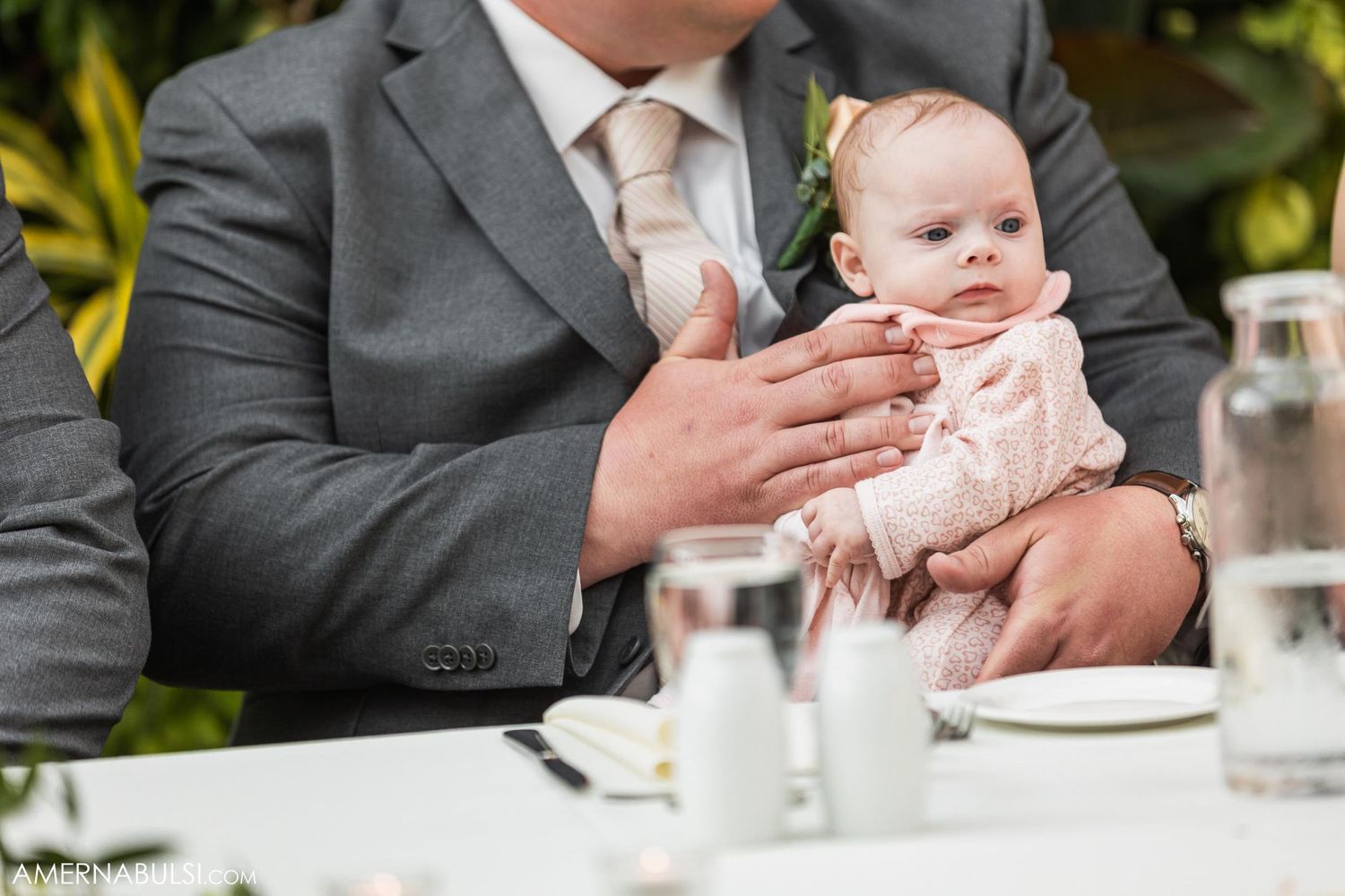 A small baby in formal wear is held during a wedding reception dinner.