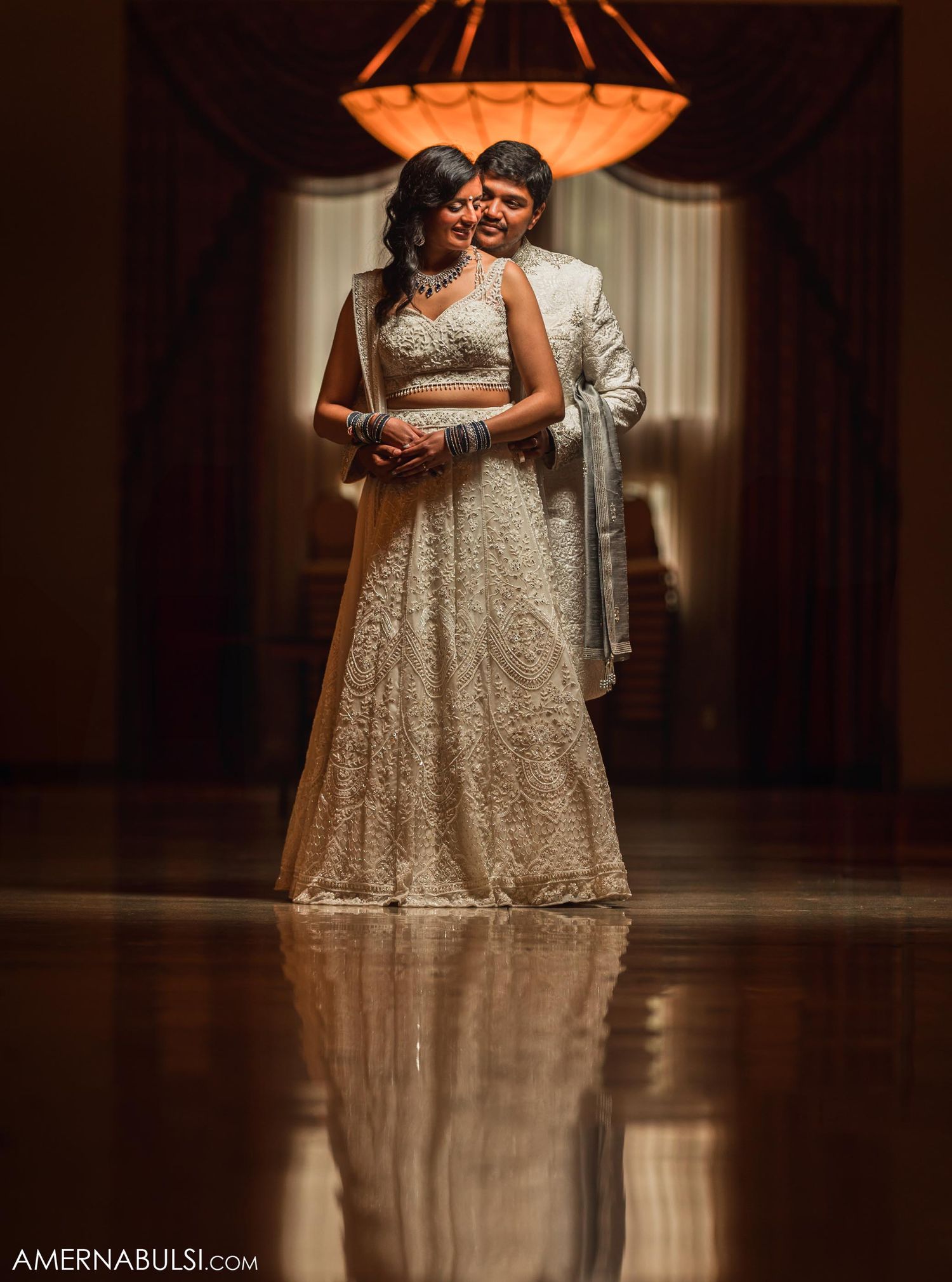 A couple poses under an ornate chandelier in a luxurious ballroom setting with reflective floors.