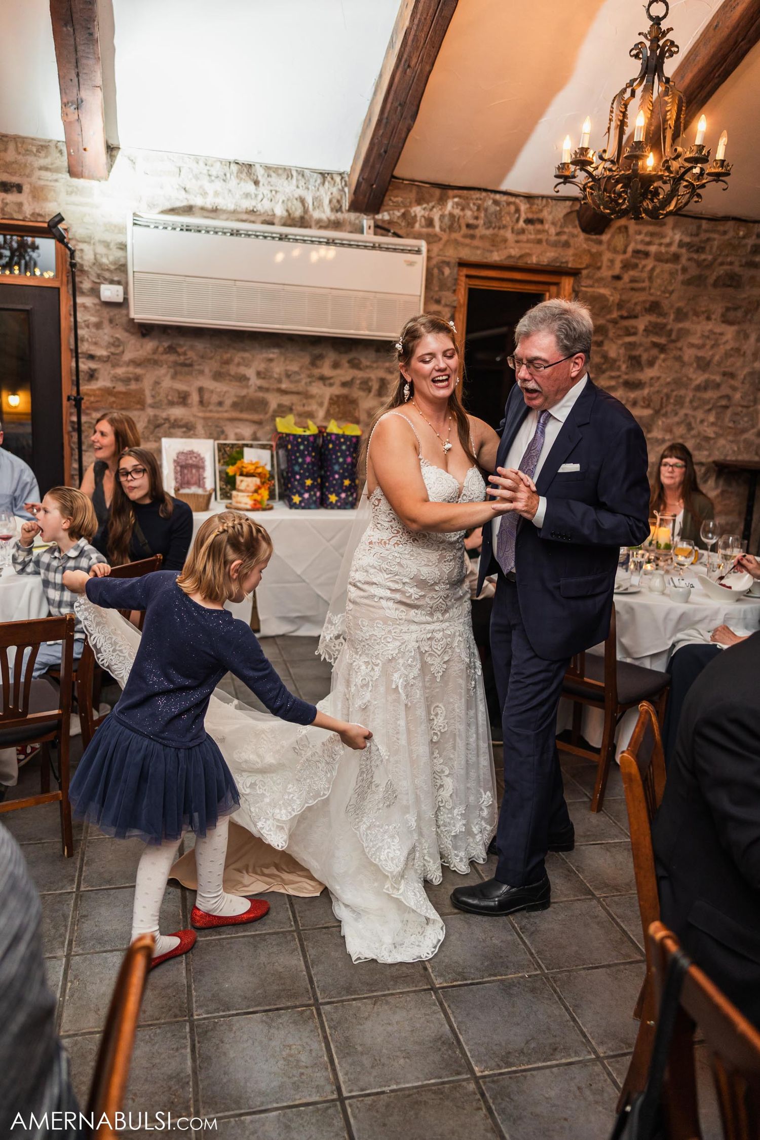 Bride in lace gown dancing with partner while young guest in navy dress twirls beside them.