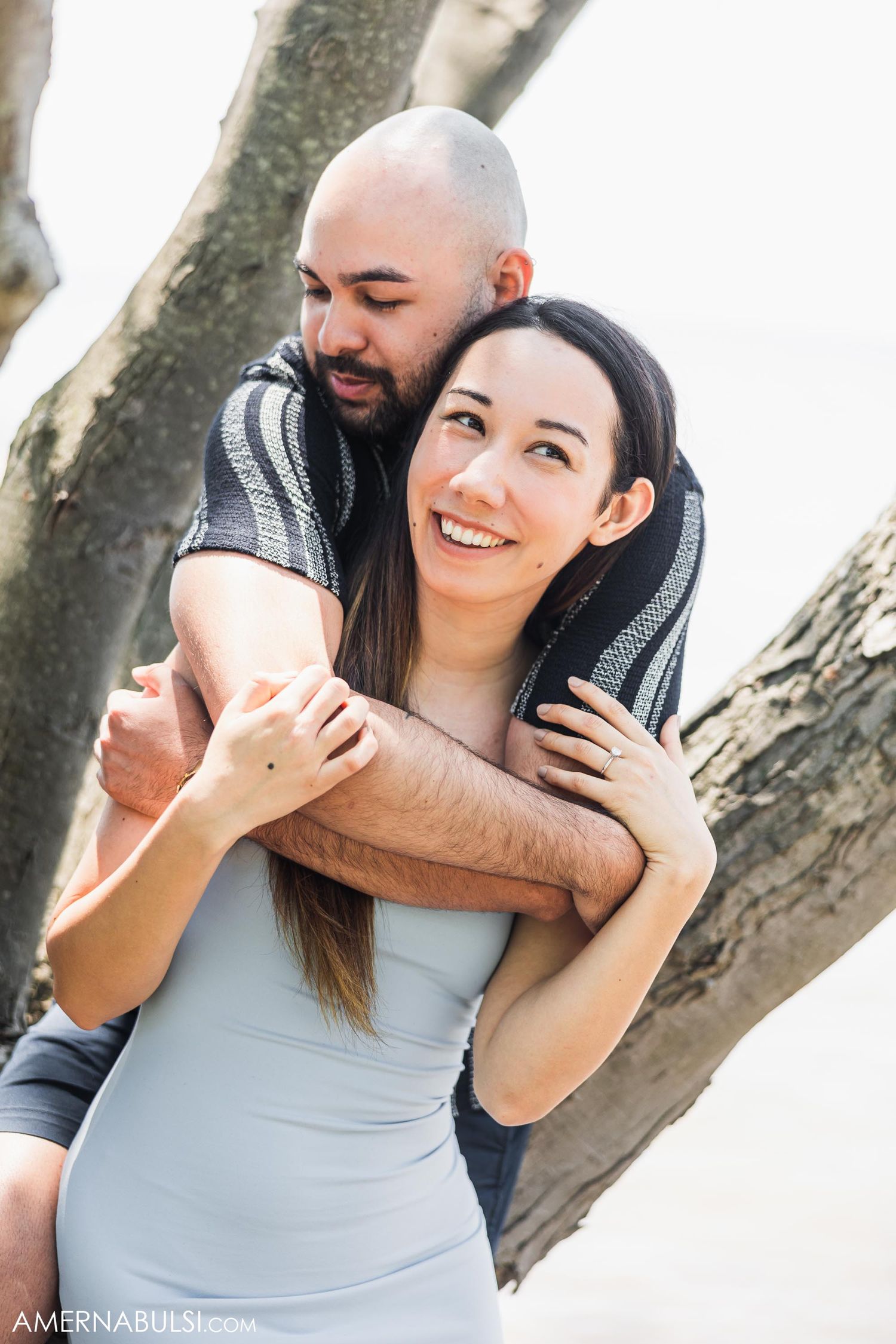 Candid outdoor portrait of a couple sharing a playful embrace near a tree.