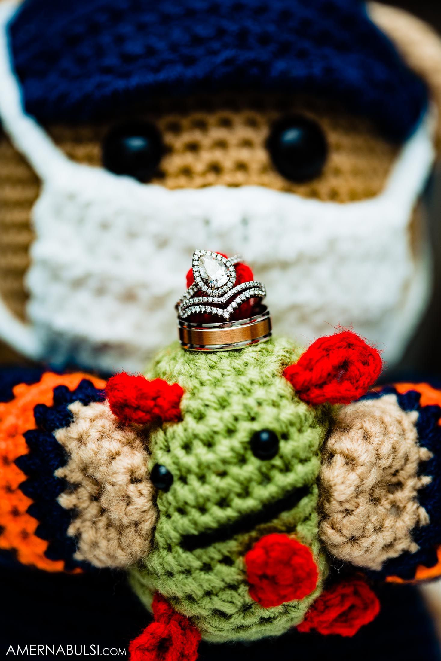 Wedding rings displayed on cute knitted plush toy with blue hat and colorful details.